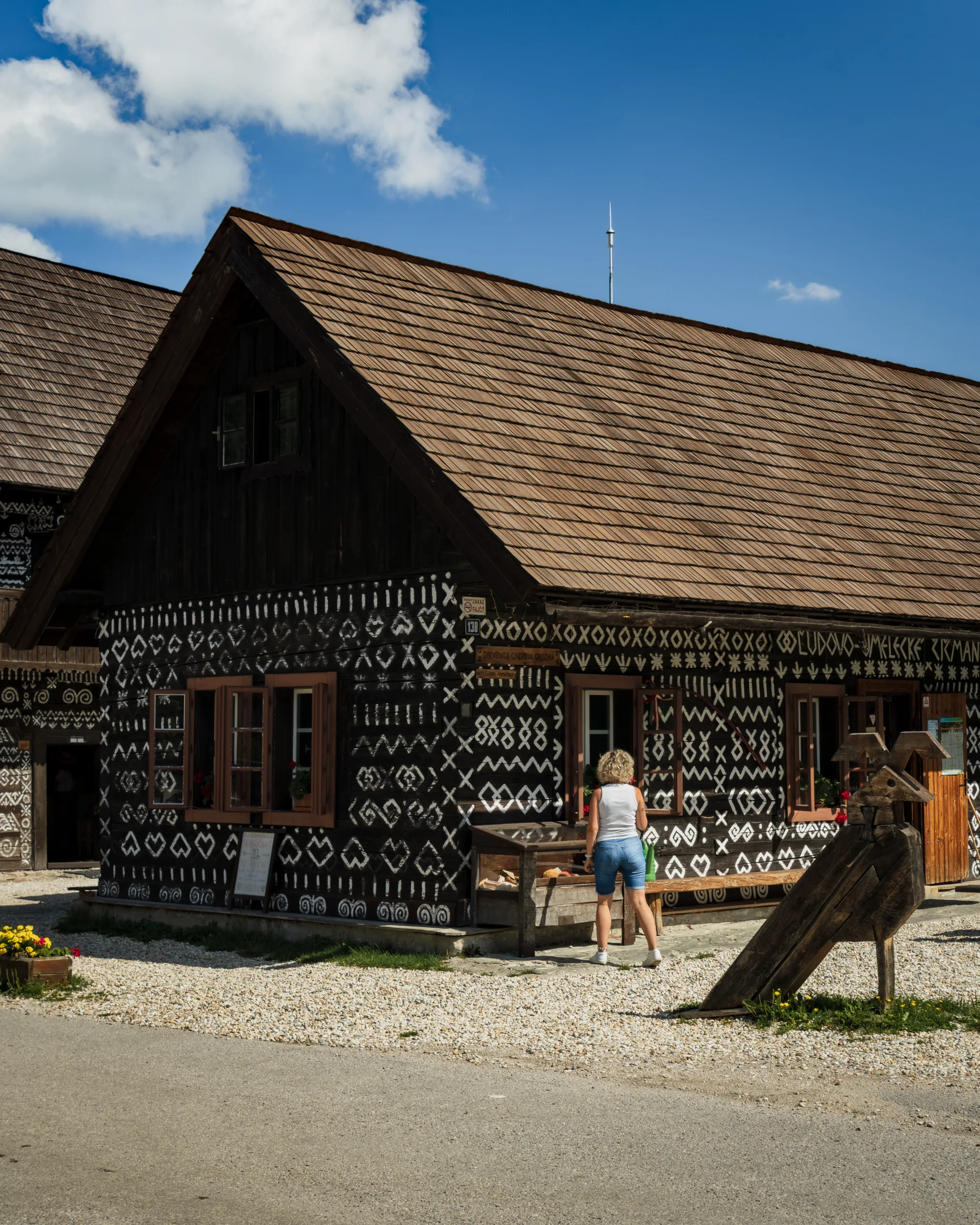 Zdobiona białymi ornamentami drewniana chata w Čičmanach na Słowacji. Przed domem stoi kobieta w letnim stroju, a obok widoczna jest drewniana rzeźba i kwiaty w donicach.