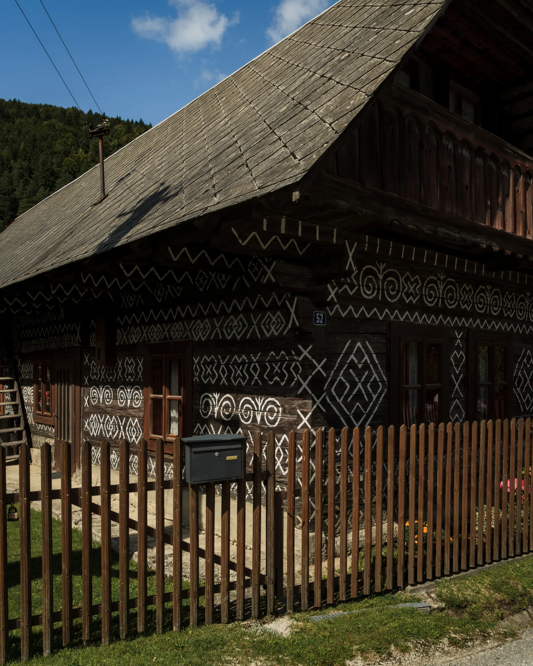 Tradycyjna drewniana chata w Čičmanach na Słowacji, zdobiona białymi geometrycznymi ornamentami na ciemnych ścianach. Przed domem znajduje się drewniany płot.