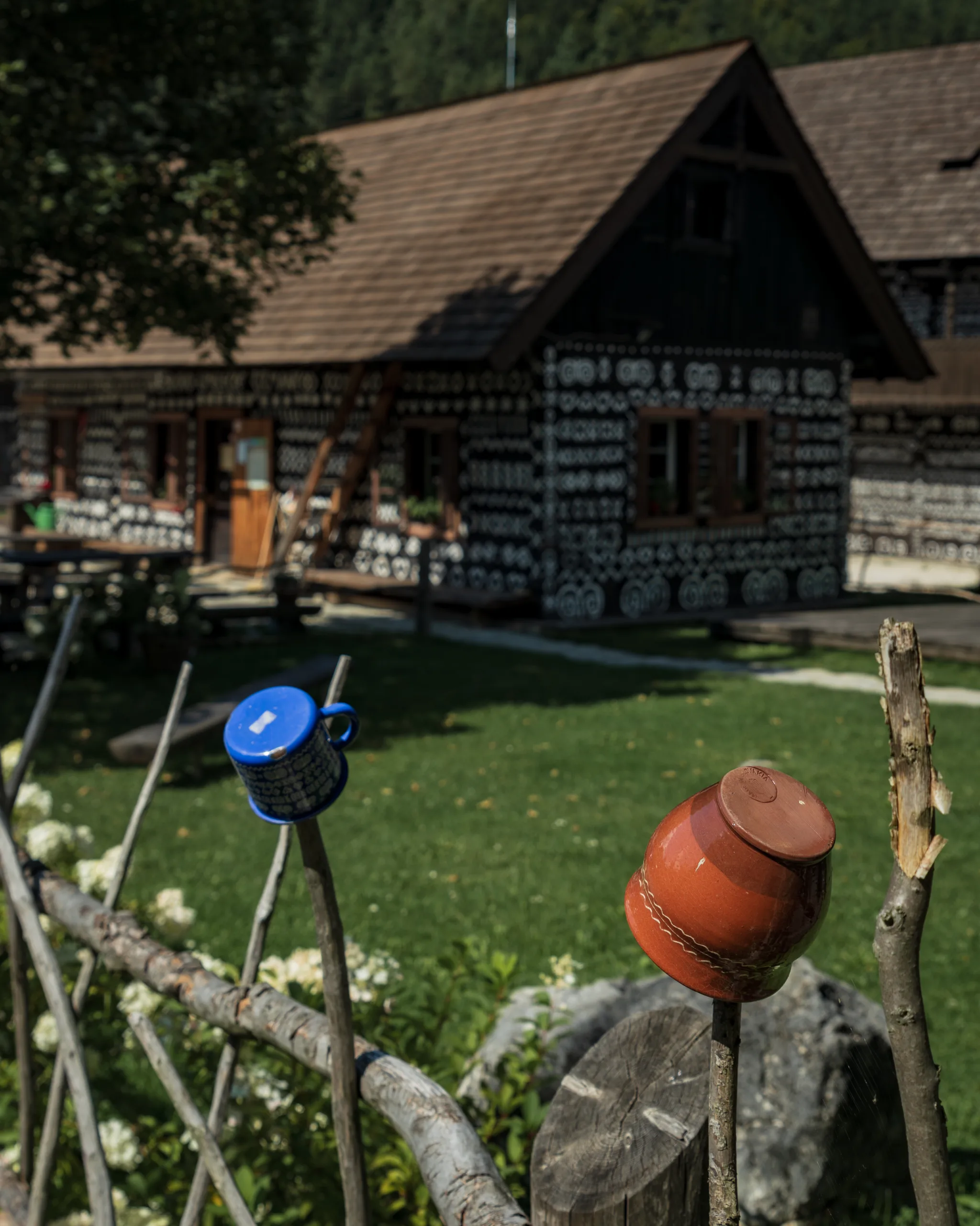 Kolorowe kubki i miski wbite na drewniane sztachety przed tradycyjną chatą w Čičmanach na Słowacji, zdobioną białymi geometrycznymi ornamentami.