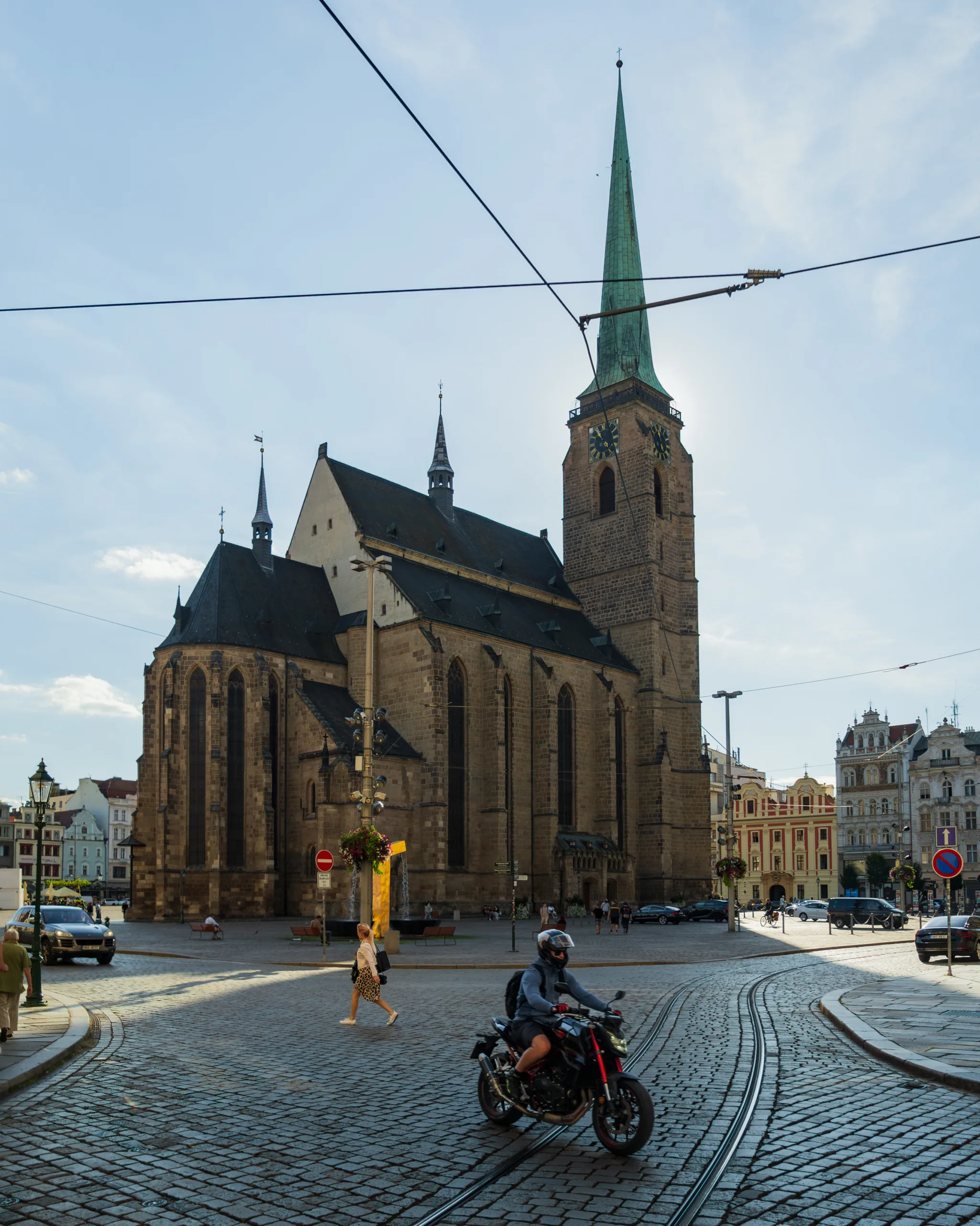 Katedra św. Bartłomieja w Pilznie – gotycka świątynia z wysoką zieloną wieżą widziana z placu, na pierwszym planie motocyklista i brukowane ulice z torami tramwajowymi.