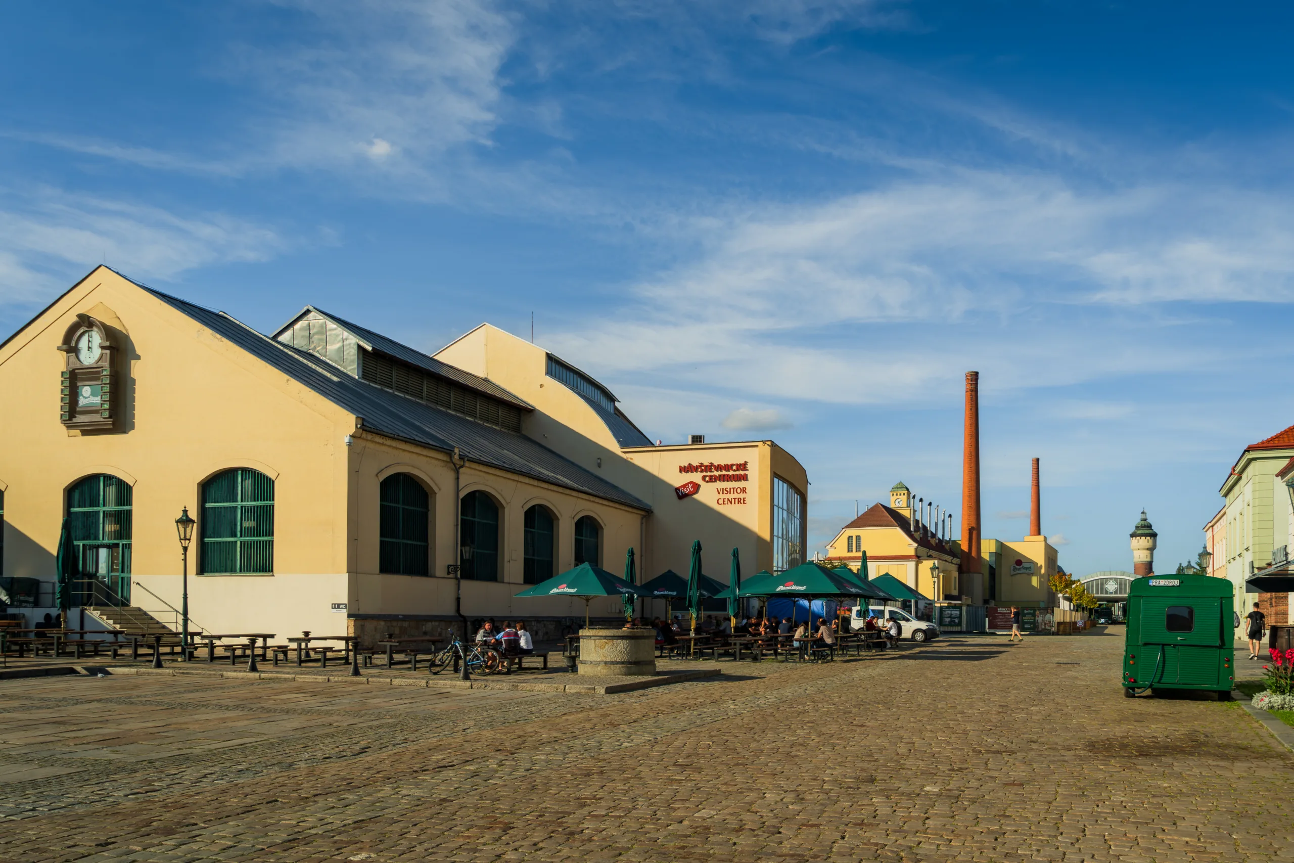 Centrum dla odwiedzających browaru Pilsner Urquell w Pilznie – żółte budynki przemysłowe z zielonymi parasolami piwnego ogródka i charakterystycznymi kominami w tle.