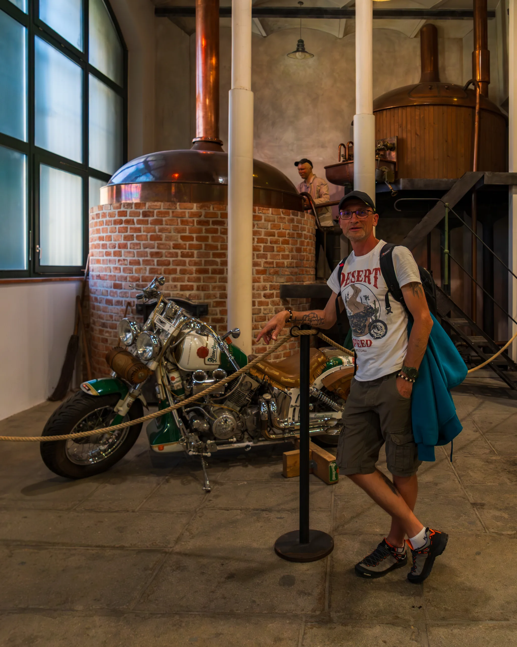Ekspozycja w browarze – miedziane kadzie warzelne, klasyczny motocykl i zwiedzający pozujący do zdjęcia.