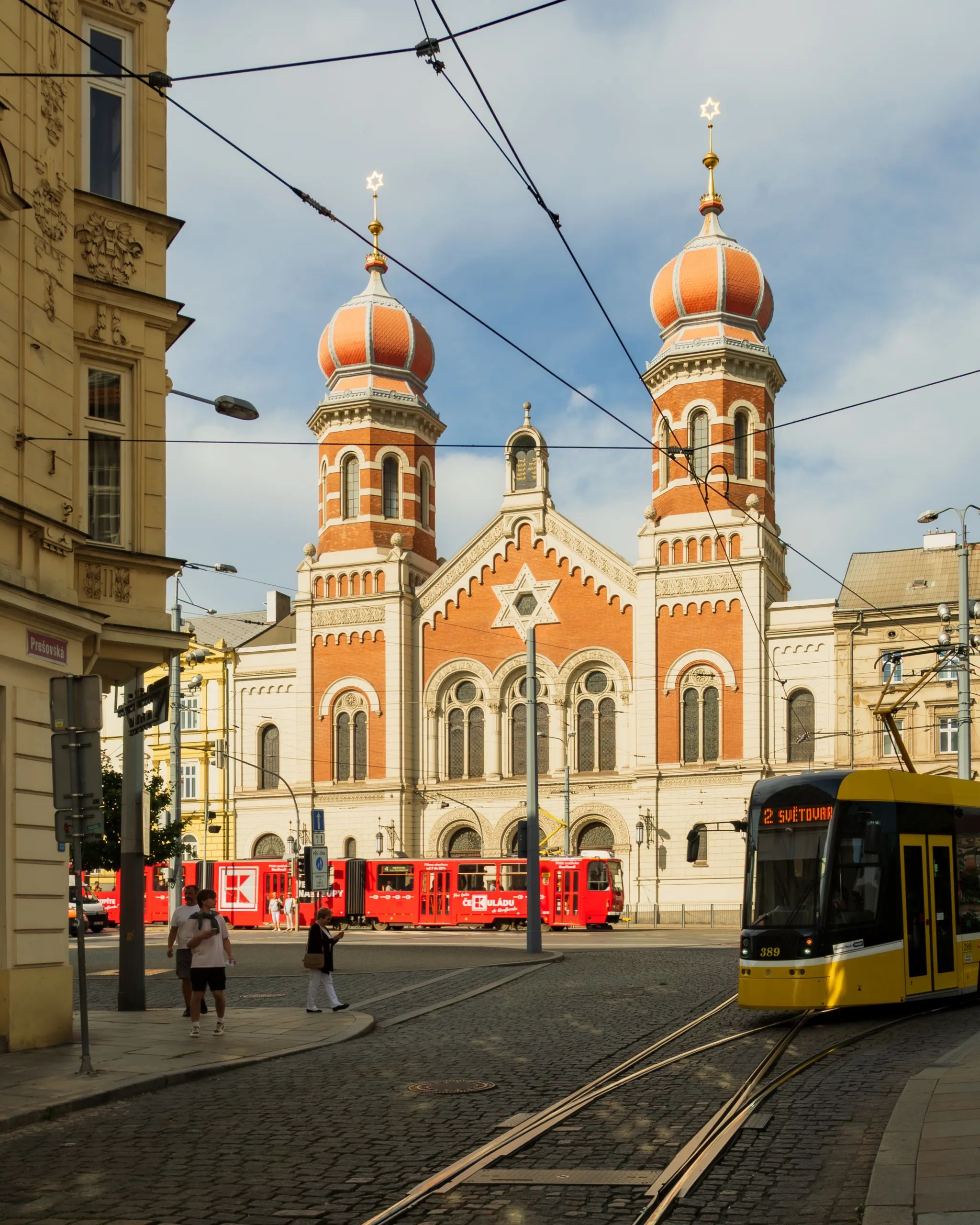 Wielka Synagoga w Pilznie – monumentalna budowla z czerwonej cegły z dwiema kopułami, widok od ulicy z przejeżdżającym żółtym tramwajem i czerwonym składem w tle.