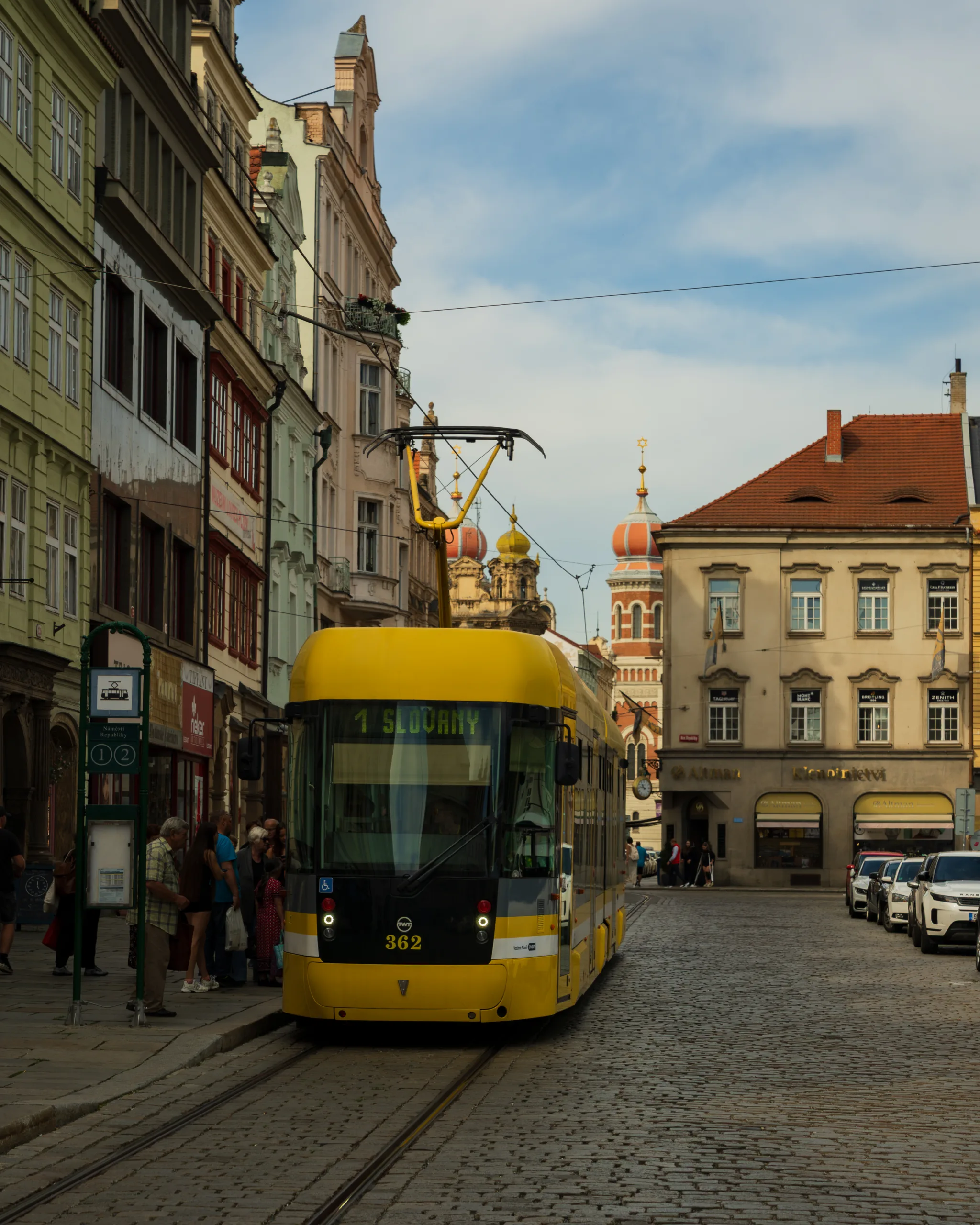 Żółty tramwaj linii nr 1 na ulicy w Pilznie, w otoczeniu kolorowych kamienic i z widokiem na synagogę z czerwonymi kopułami w tle.