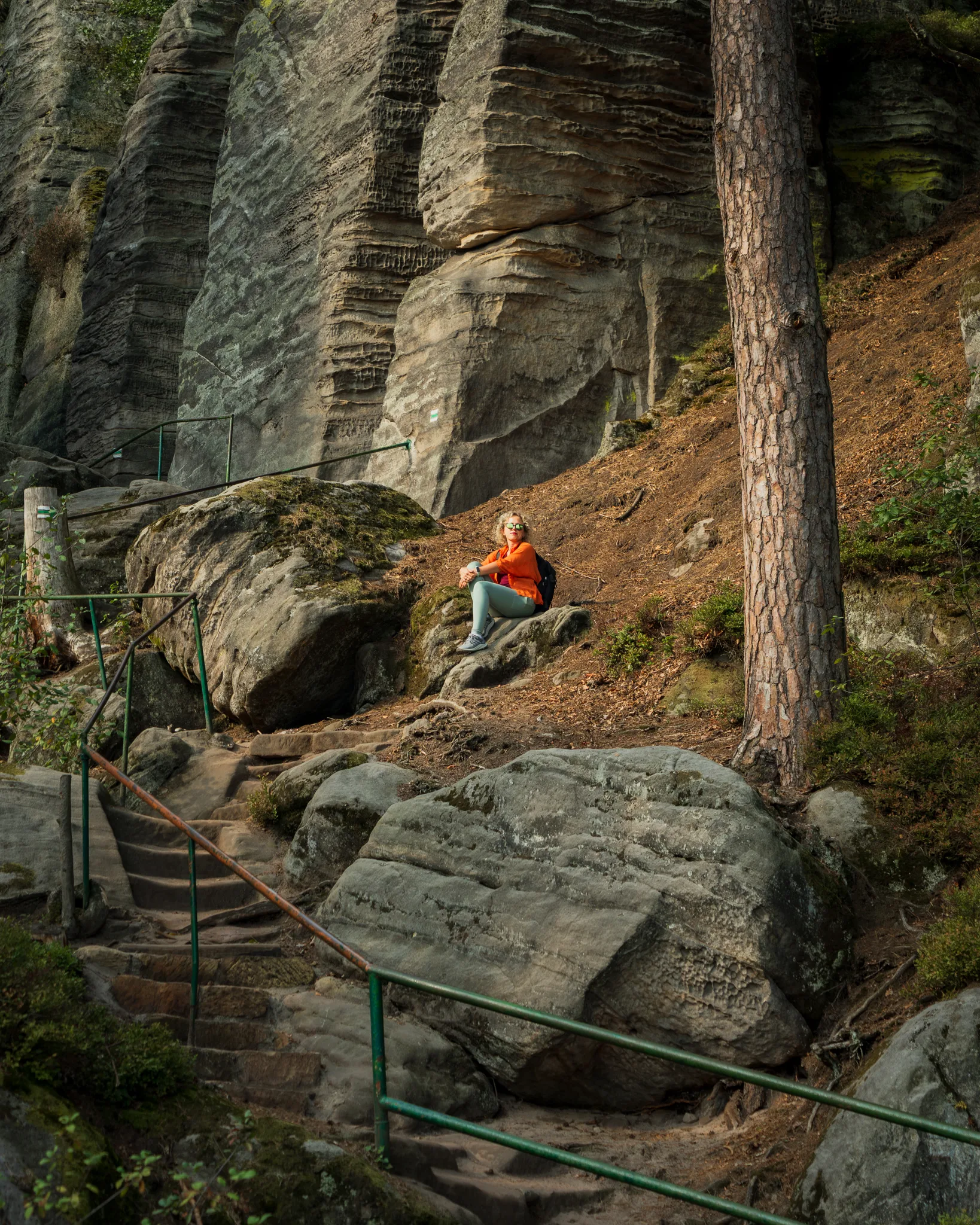 Turystka w pomarańczowej kurtce odpoczywająca na skale w Czeskich Skałach Adrszpaskich, otoczona stromymi ścianami skalnymi i lasem.