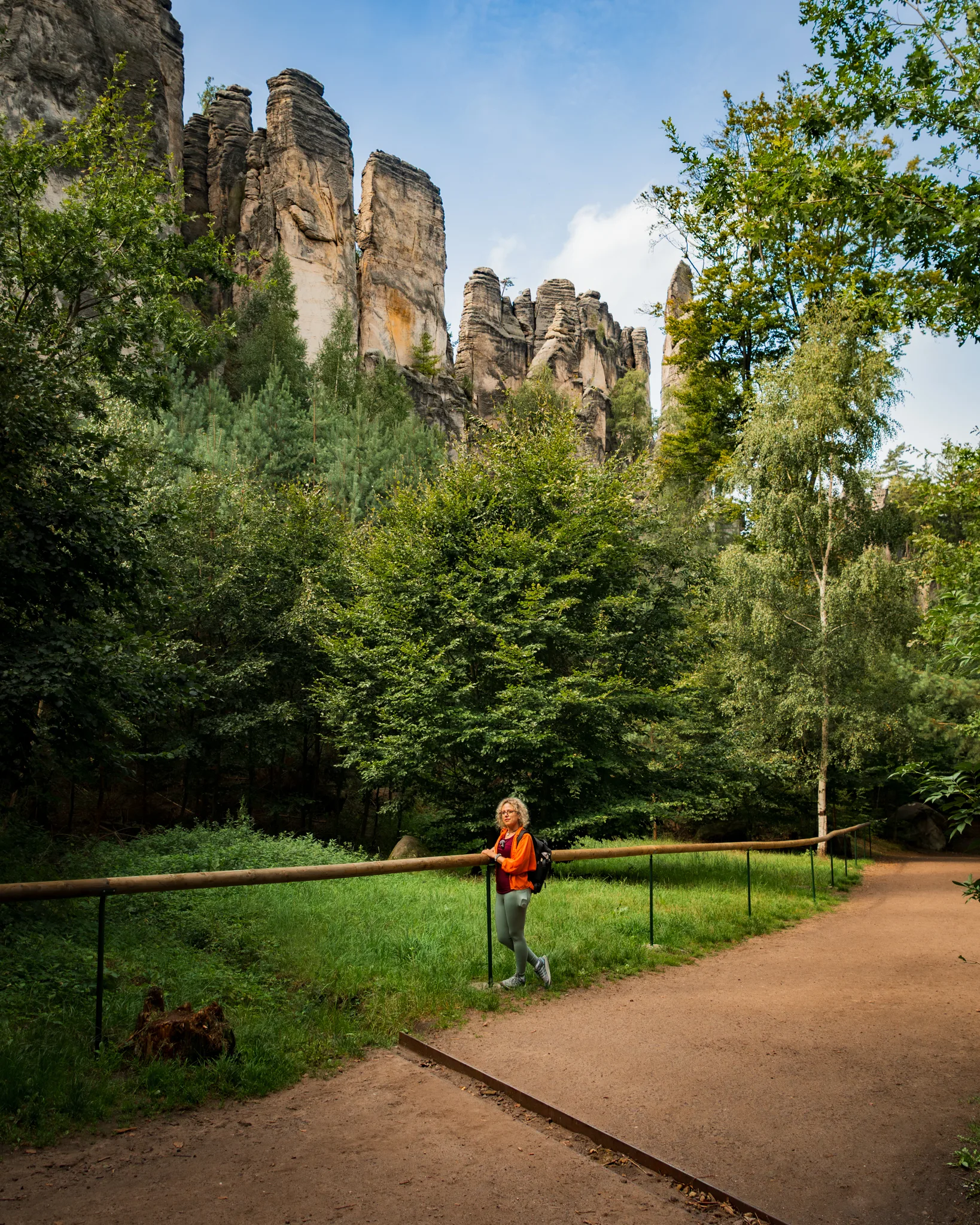 Kobieta na szlaku wśród drzew, w tle monumentalne formacje skalne w Czeskiej Szwajcarii.