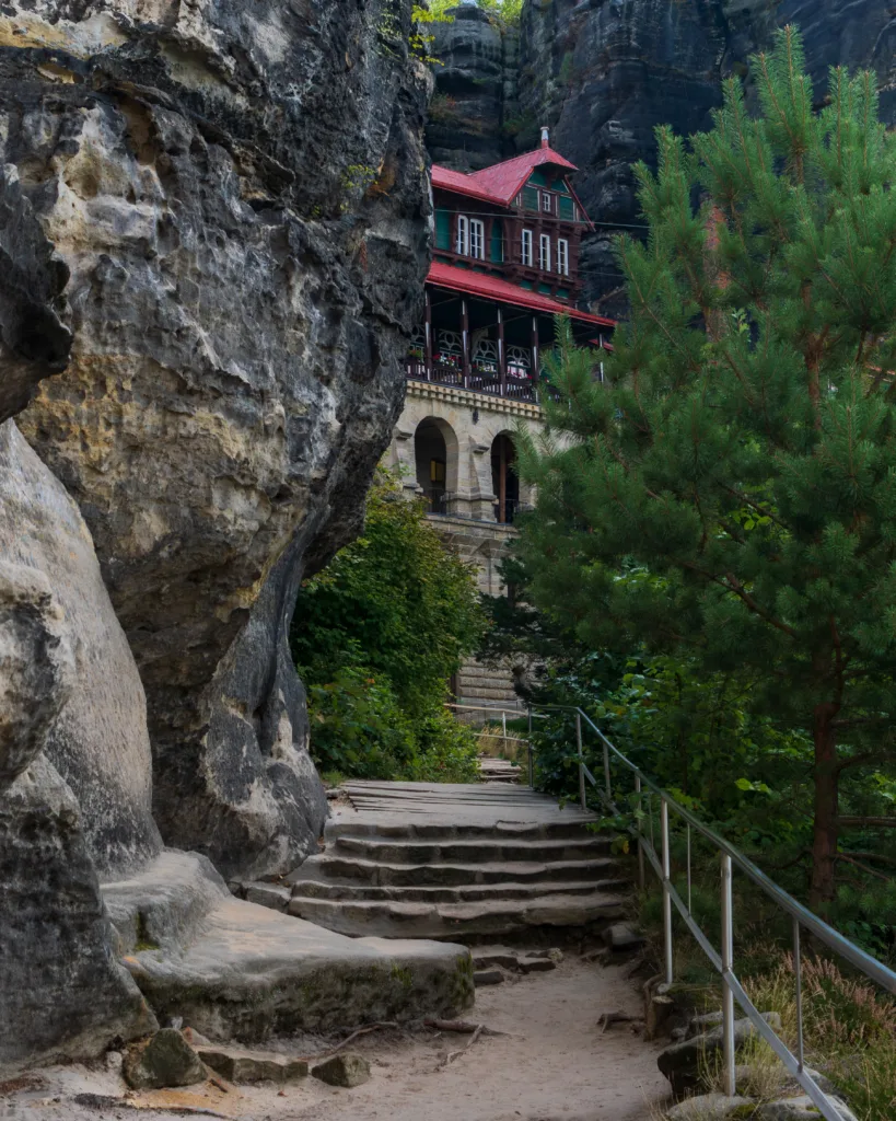 Wejście do schroniska Sokolí hnízdo pod Pravčicką Bramą w Czeskiej Szwajcarii, widziane od kamiennych schodów wśród skał i drzew.