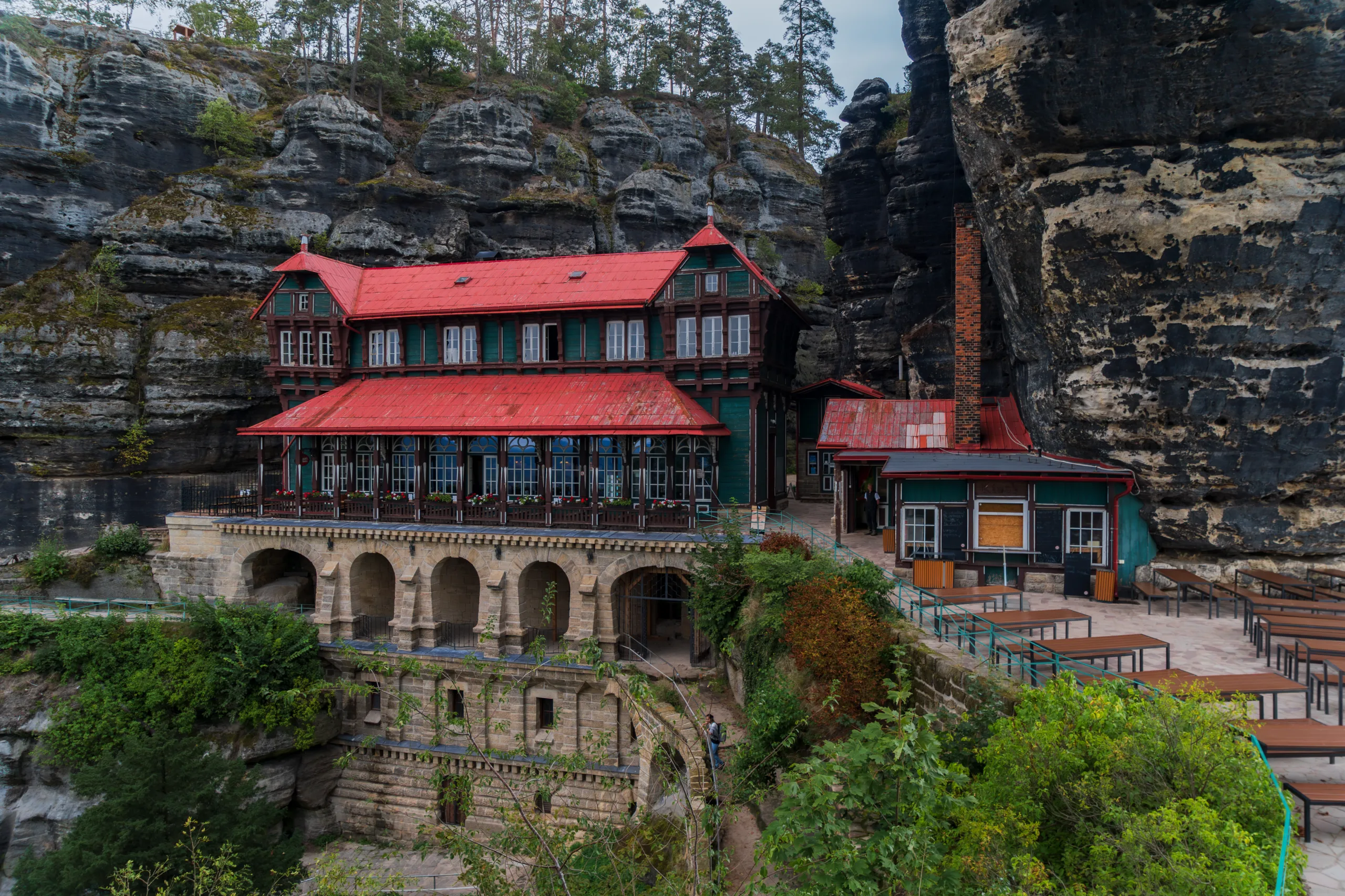 Schronisko Sokolí hnízdo z czerwonym dachem, zbudowane przy Pravčickiej Bramie w Czeskiej Szwajcarii, otoczone monumentalnymi skałami.