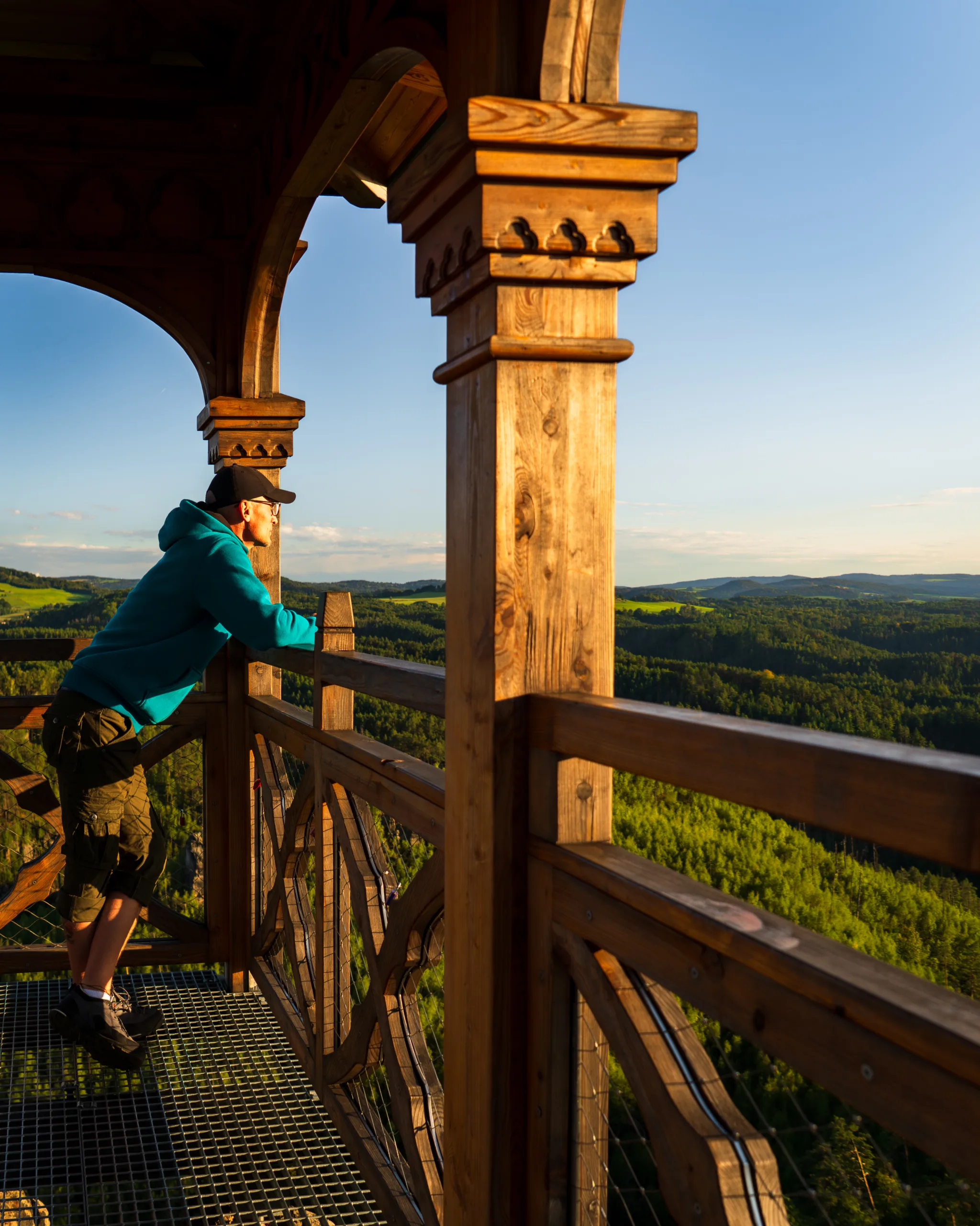 Turysta podziwiający panoramę zielonych wzgórz z drewnianej wieży widokowej o zachodzie słońca.