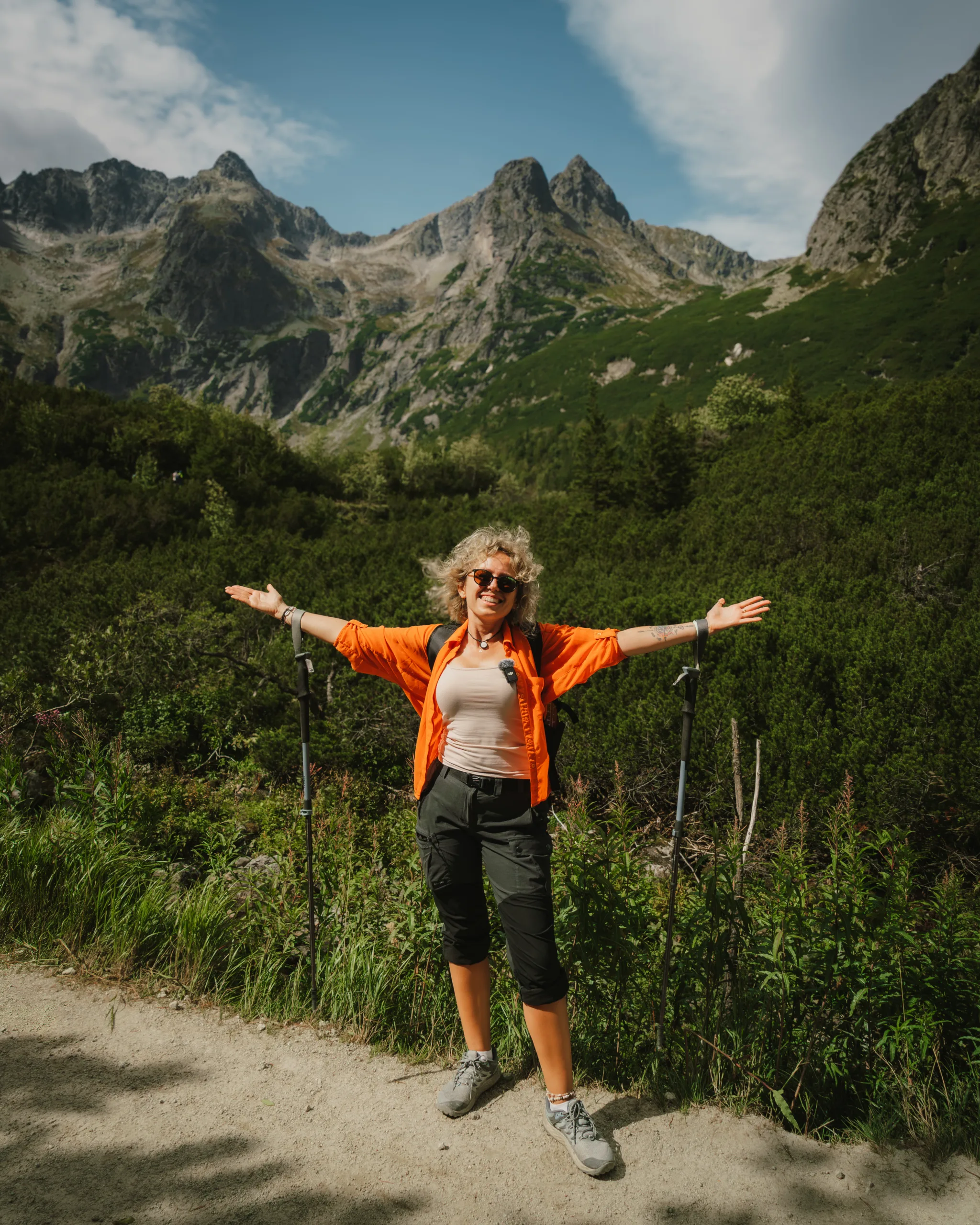 Turystka w pomarańczowej koszuli z rozłożonymi rękami na szlaku w Tatrach Wysokich, w otoczeniu gór i zieleni.