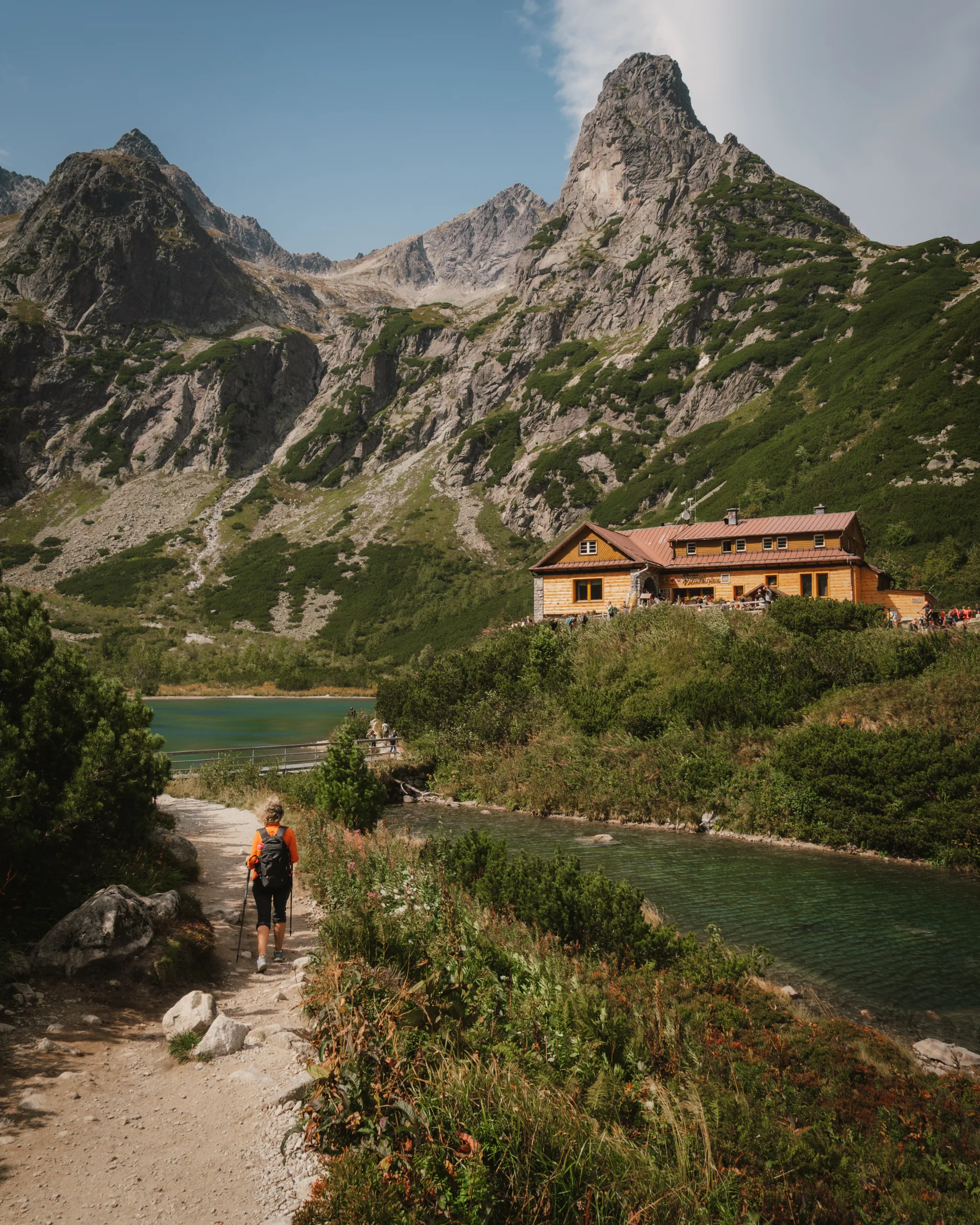 Turysta idący górskim szlakiem nad jeziorem w Tatrach Wysokich, obok schroniska położonego u stóp skalistych szczytów.