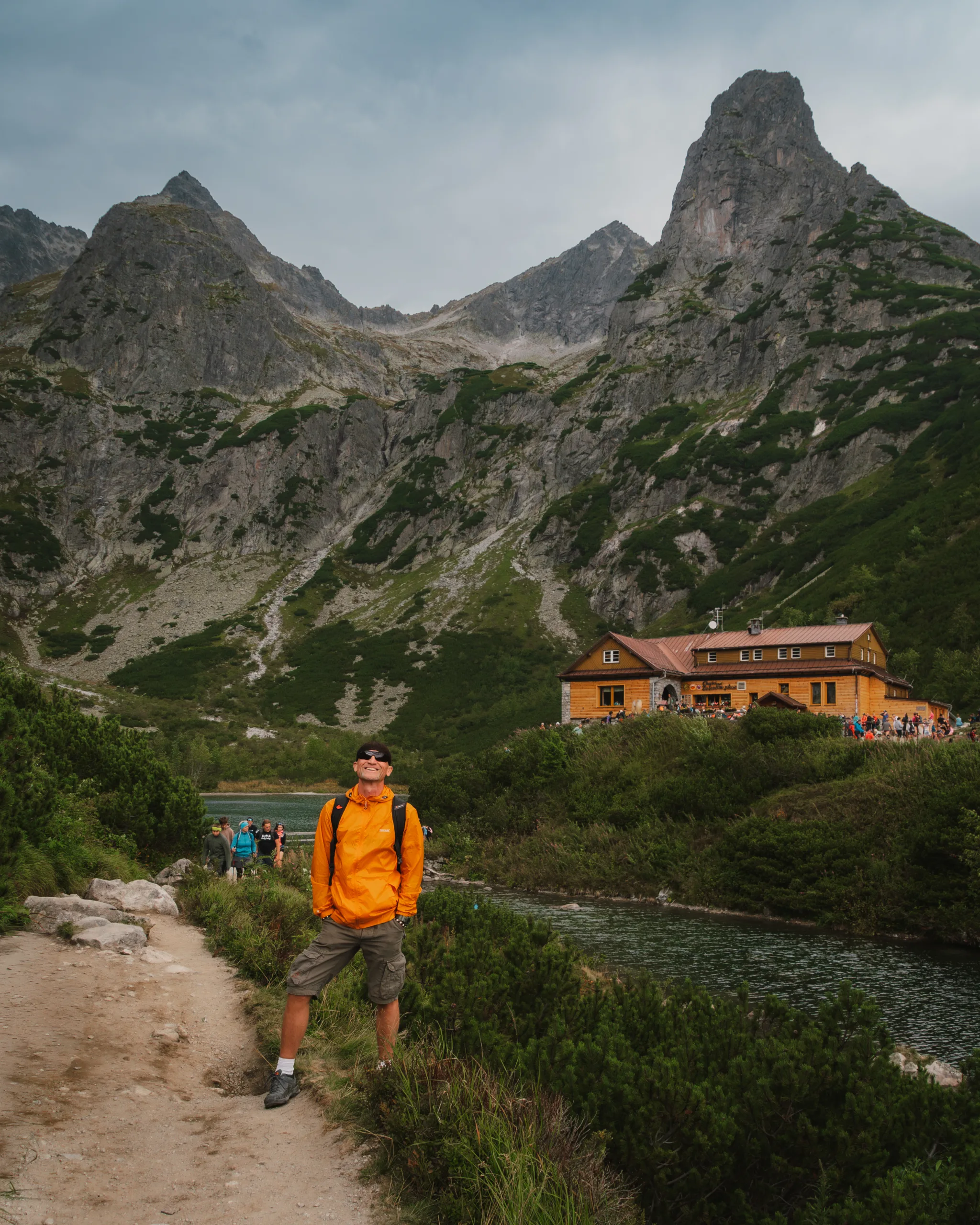 Turysta w pomarańczowej kurtce na szlaku w Tatrach Wysokich, z widokiem na schronisko i skaliste szczyty w tle.