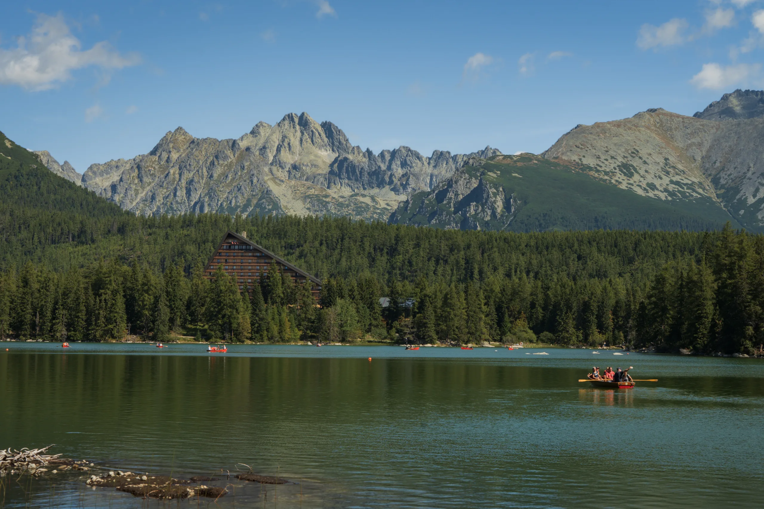 Štrbskie Jezioro w Tatrach Wysokich z hotelem Patria i górami w tle, spokojna tafla wody i czerwone łódki.