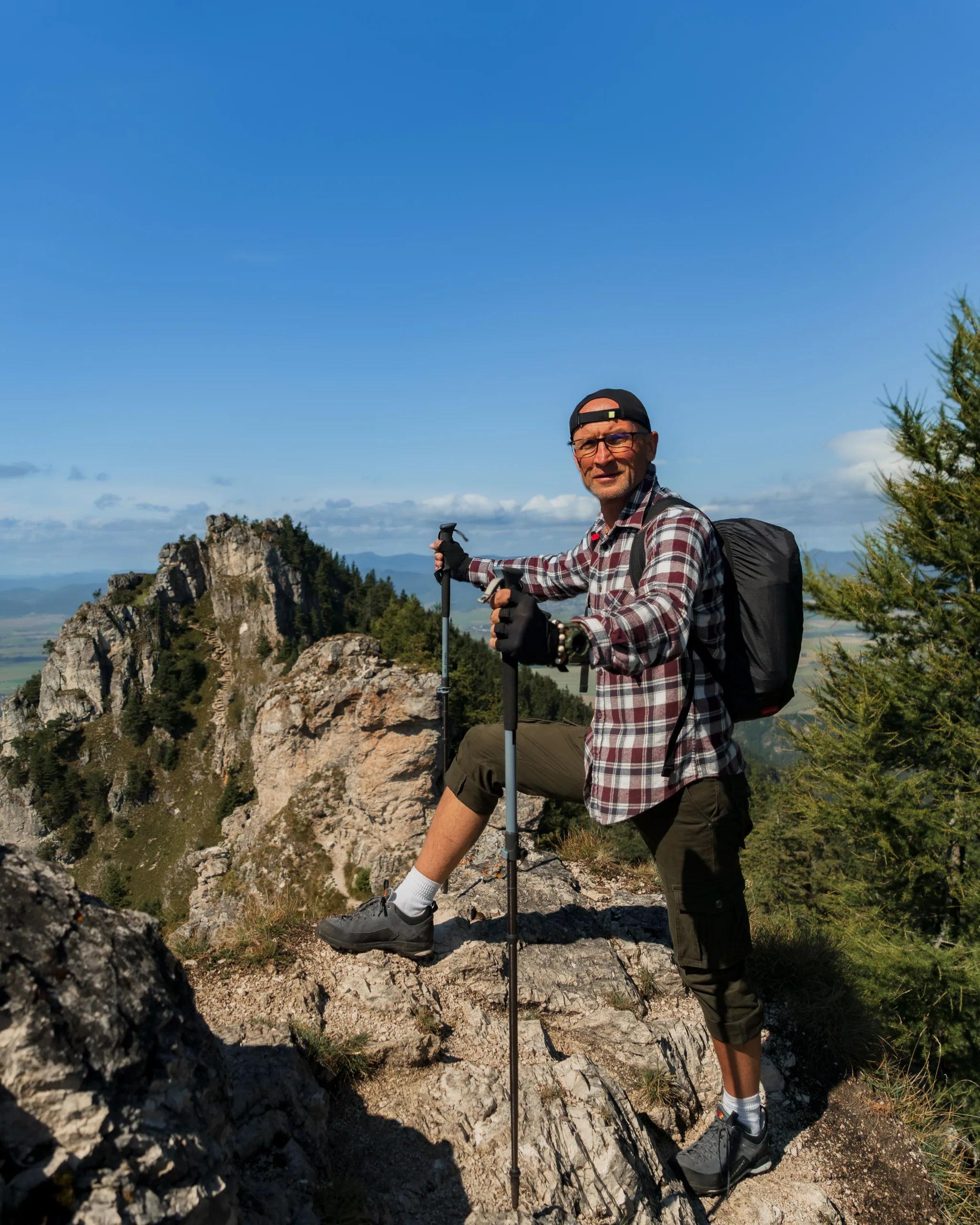 Turysta w koszuli w kratę z plecakiem i kijkami trekkingowymi na szczycie góry w Wielkiej Fatrze, z panoramą skalistego grzbietu w tle.