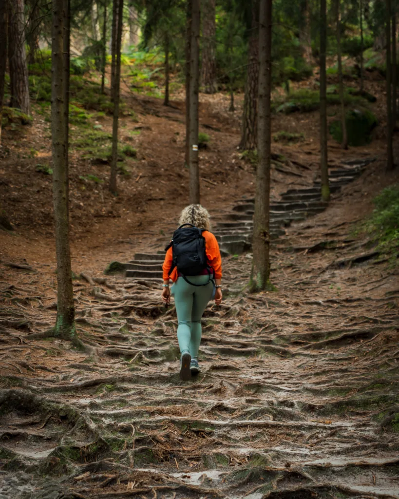 Turystka z plecakiem idąca leśnym szlakiem w Czeskim Raju, otoczonym drzewami i korzeniami na ścieżce.