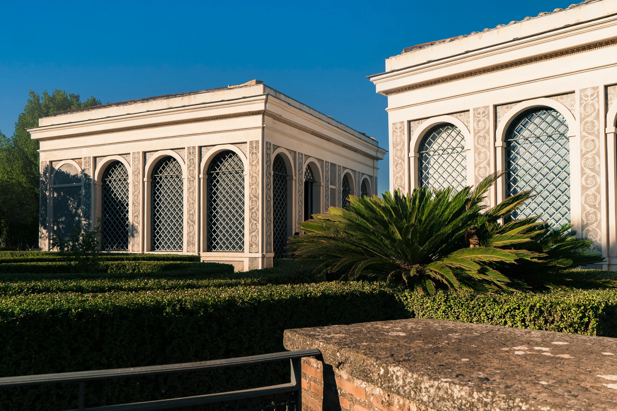 Eleganckie pawilony ogrodowe na Palatynie w Rzymie, z geometrycznymi żywopłotami i palmą w popołudniowym słońcu.