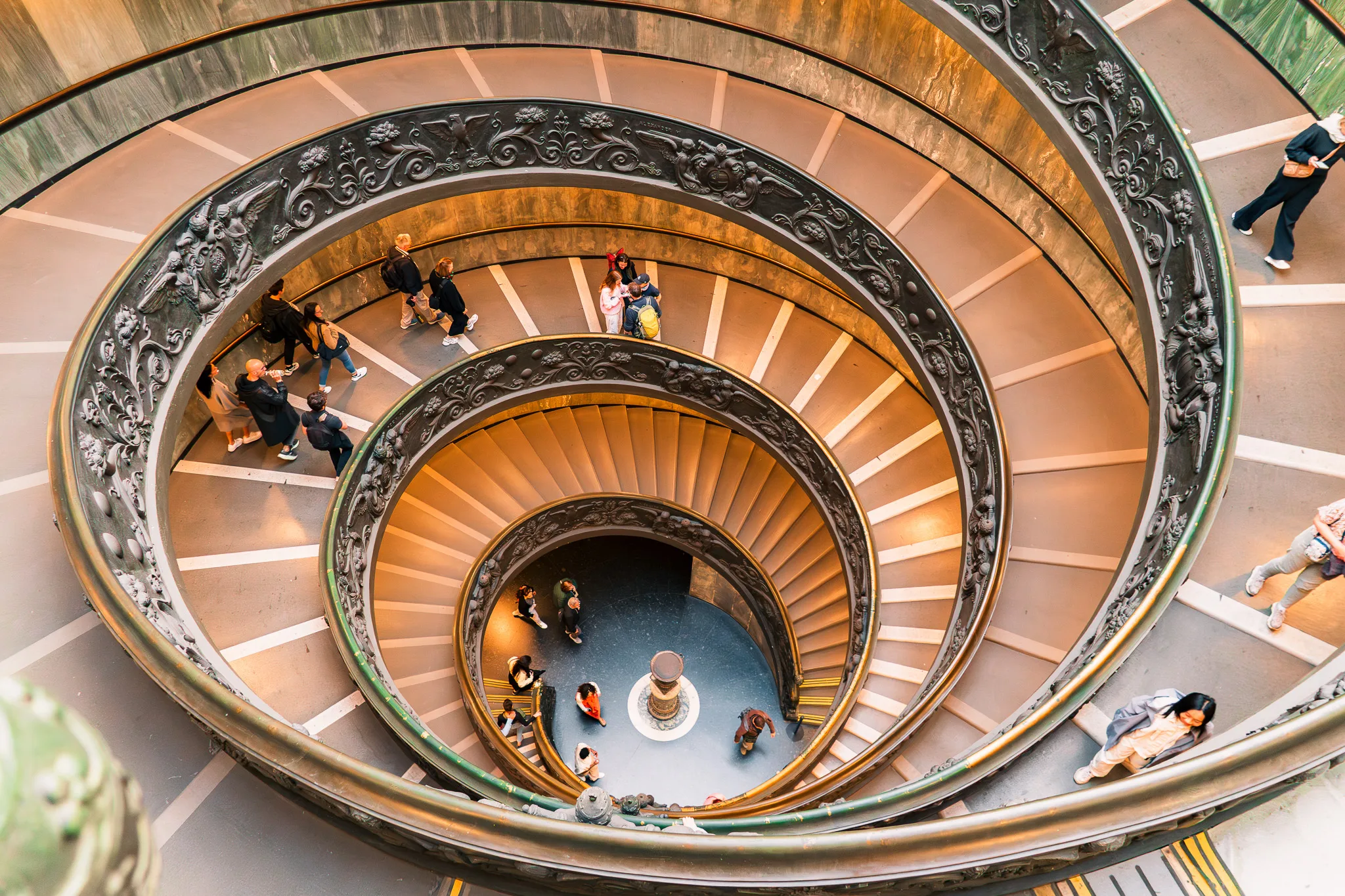 Spiralne schody w Muzeach Watykańskich, ozdobione bogato rzeźbioną balustradą i oświetlone ciepłym światłem, widziane z góry.