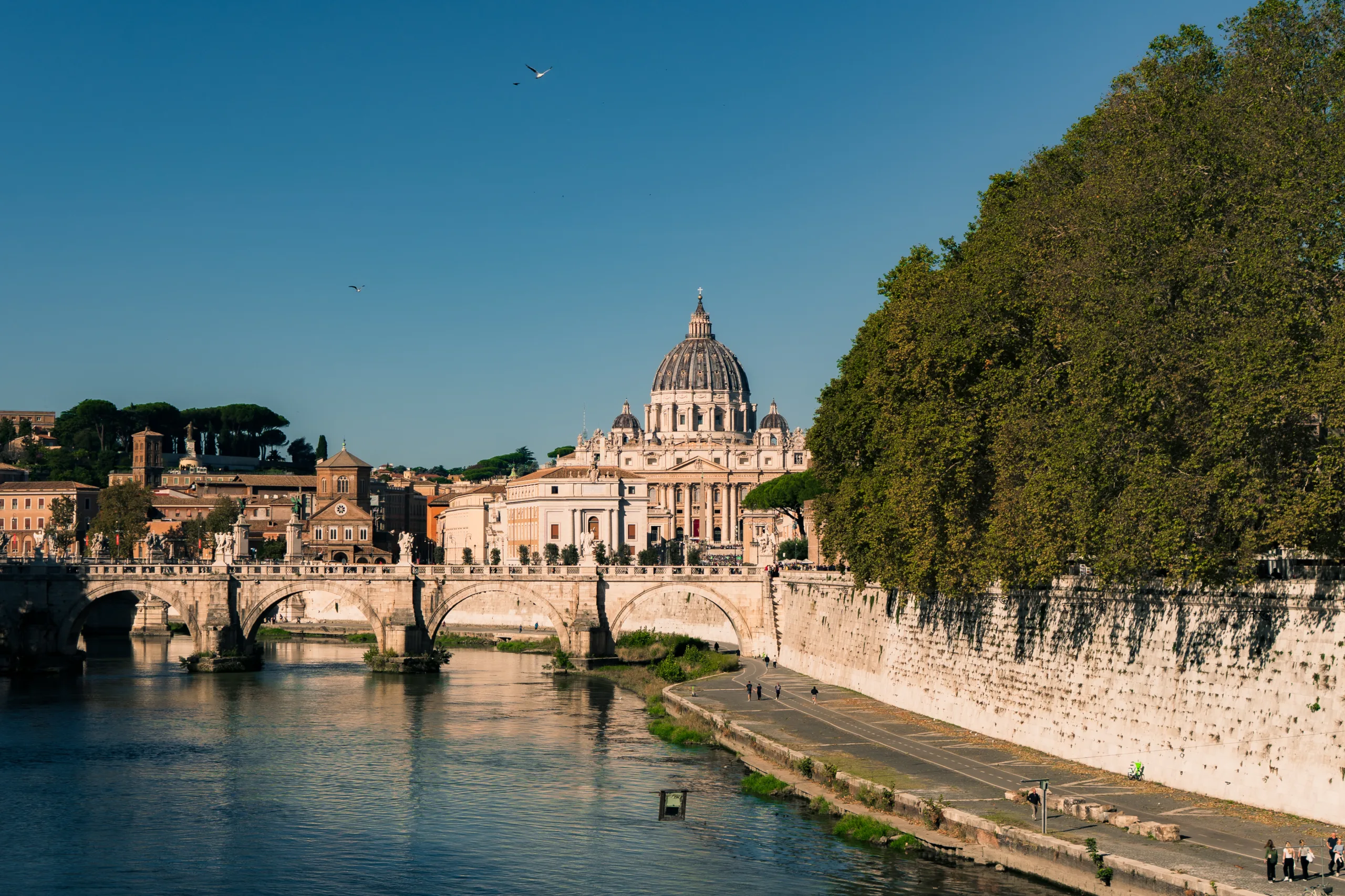 Widok na Bazylikę św. Piotra w Watykanie z mostem Sant’Angelo i rzeką Tyber w Rzymie, w popołudniowym słońcu.