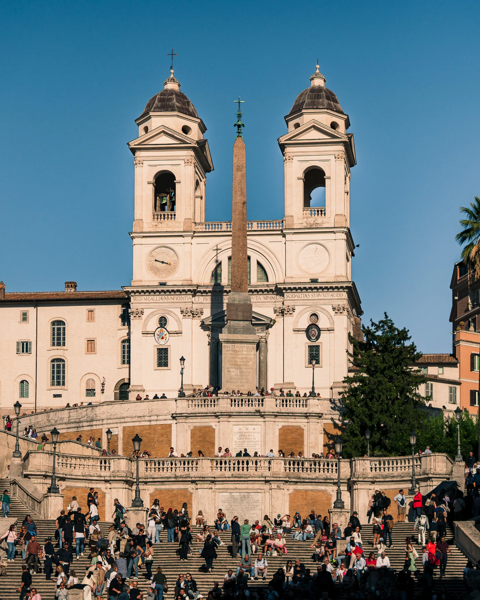 Kościół Trinità dei Monti i Schody Hiszpańskie w Rzymie w słoneczny dzień, pełne turystów odpoczywających na schodach.
