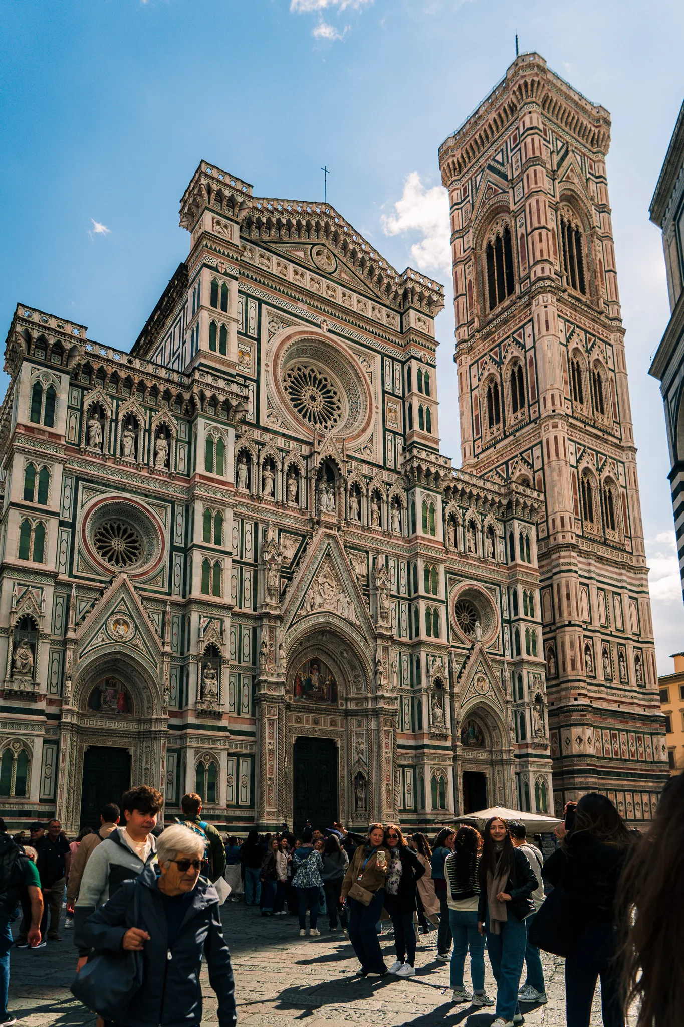 Katedra Santa Maria del Fiore we Florencji z fasadą z białego, zielonego i różowego marmuru oraz dzwonnicą Giotta, przed którą spacerują turyści.