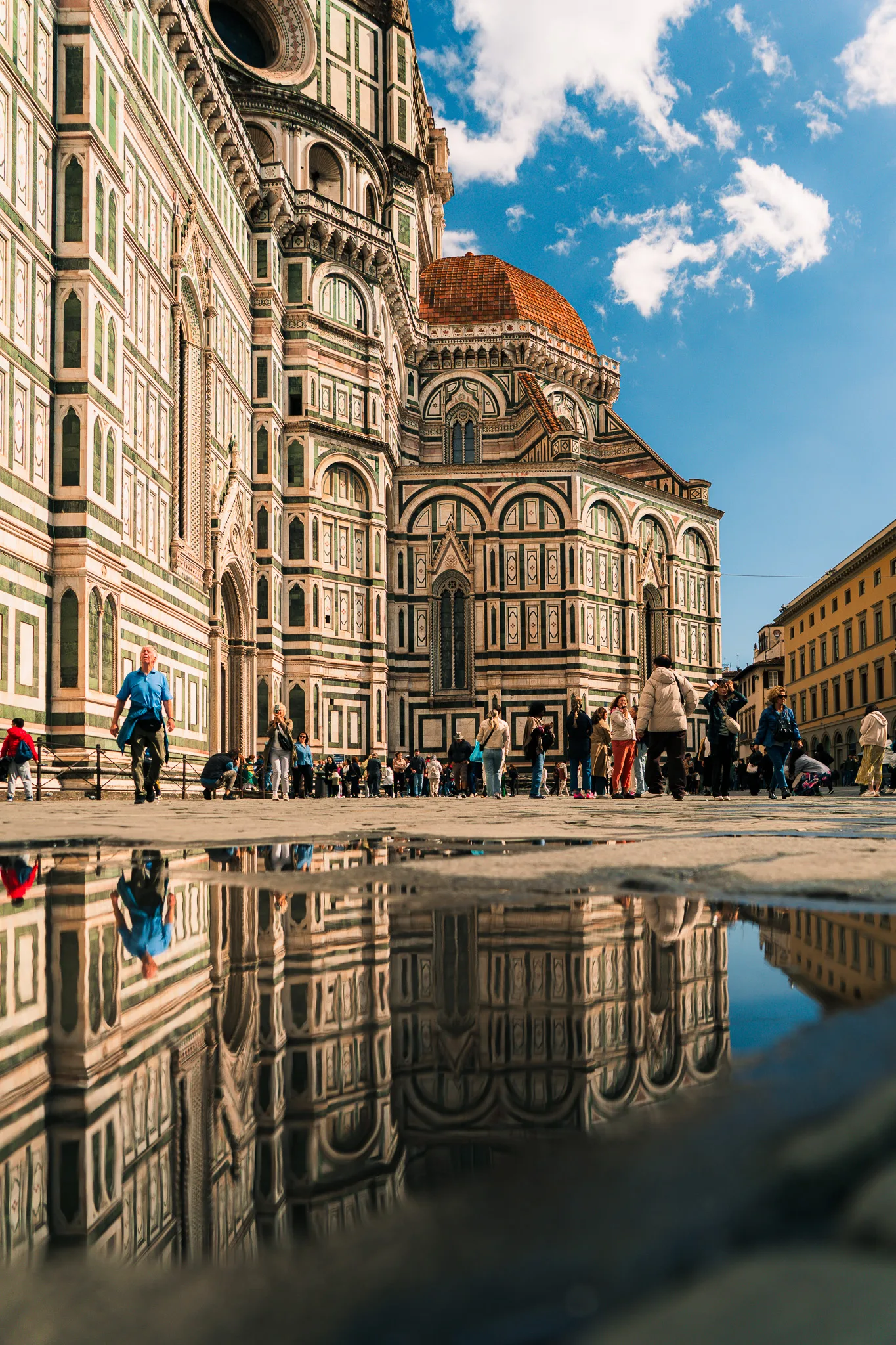 Katedra Santa Maria del Fiore we Florencji z odbiciem w kałuży i turystami spacerującymi po Piazza del Duomo w słoneczny dzień.