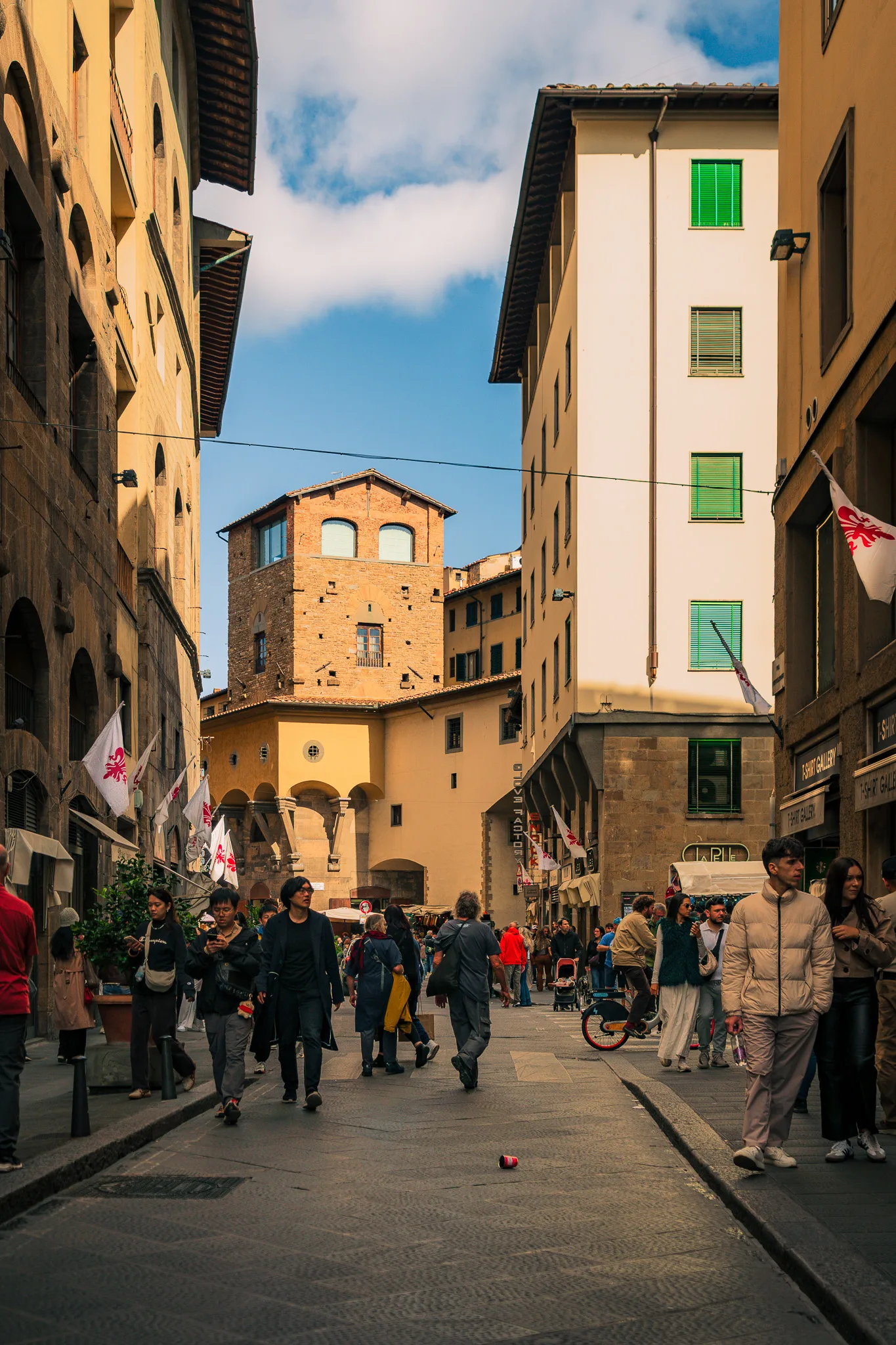 Ulica we Florencji pełna spacerujących ludzi, z widokiem na kamienice i średniowieczną wieżę w tle, w pobliżu Ponte Vecchio.