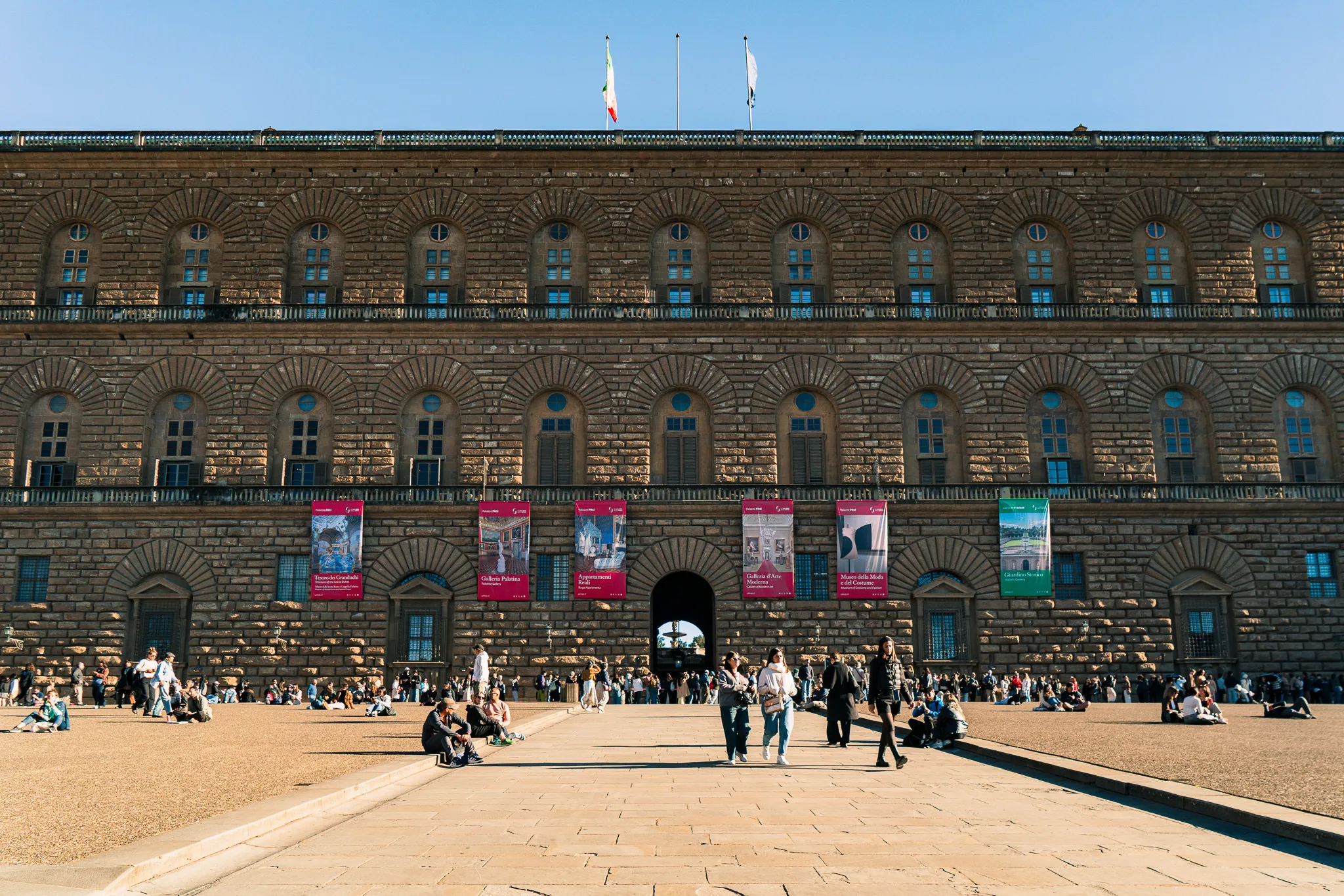 Pałac Pitti we Florencji z szerokim dziedzińcem pełnym turystów odpoczywających w słońcu.