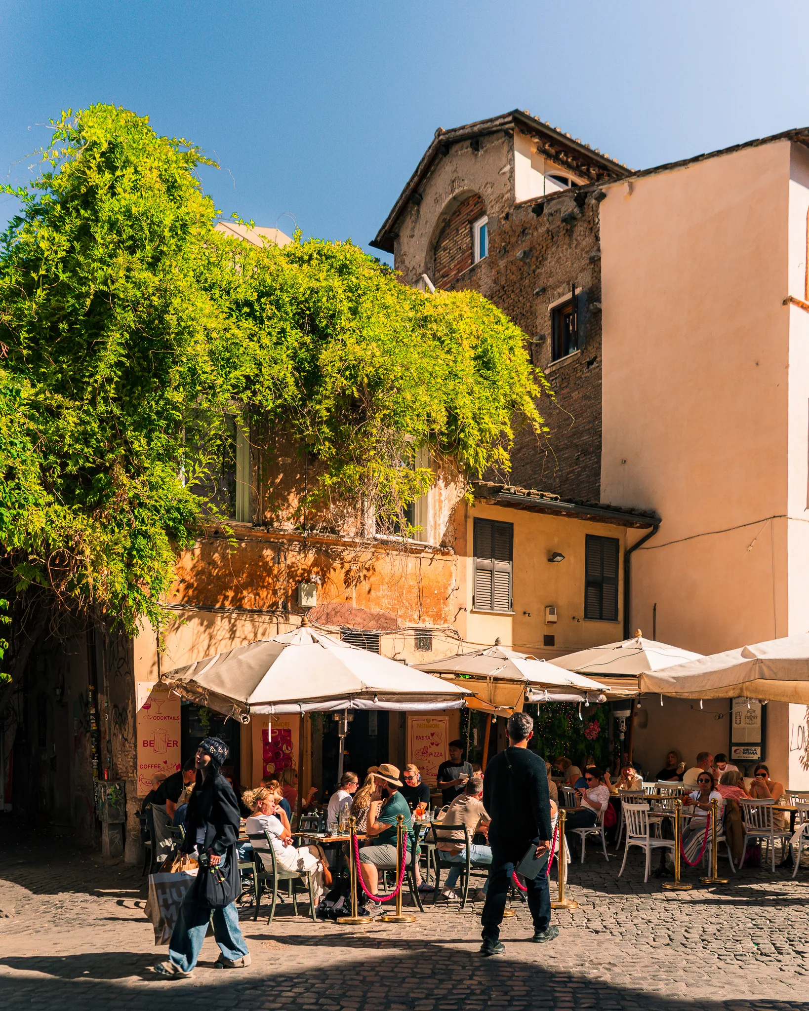 Klimatyczna kawiarnia w dzielnicy Trastevere w Rzymie z ludźmi siedzącymi przy stolikach w cieniu winorośli i starych kamienic.