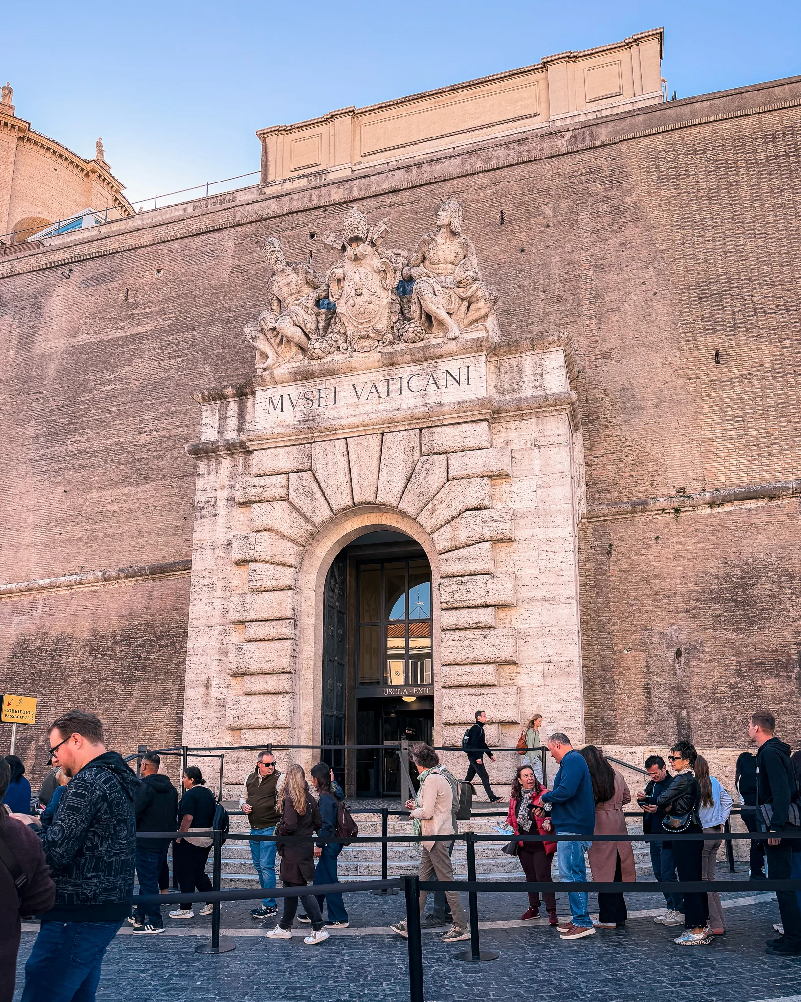 Wejście do Muzeów Watykańskich w Watykanie z napisem Musei Vaticani i turystami stojącymi w kolejce przed monumentalną bramą.