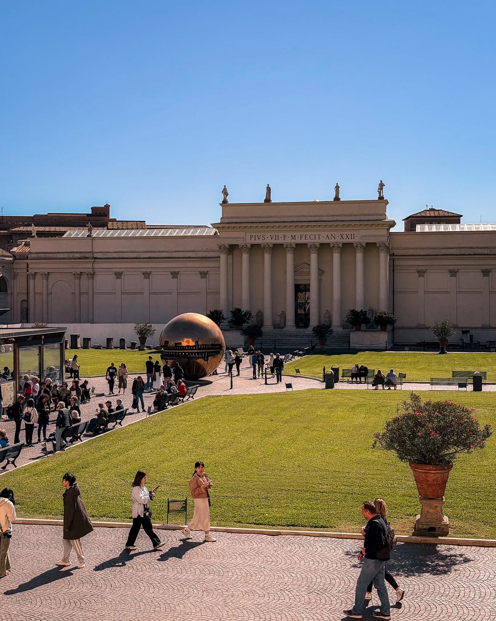 Dziedziniec Pigna w Muzeach Watykańskich z rzeźbą złotej kuli 'Sfera con Sfera' i turystami spacerującymi po zielonym trawniku.