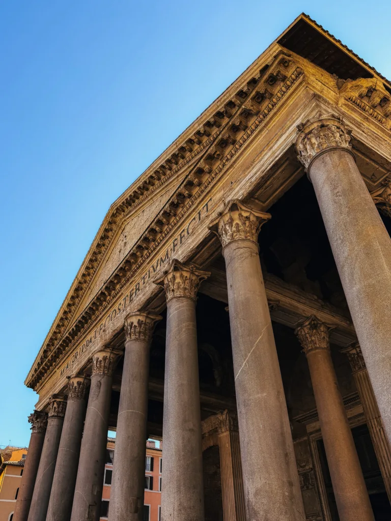 Kolumnowy portyk Panteonu w Rzymie z napisem dedykacyjnym Agrypy, ukazany z perspektywy od dołu na tle błękitnego nieba.