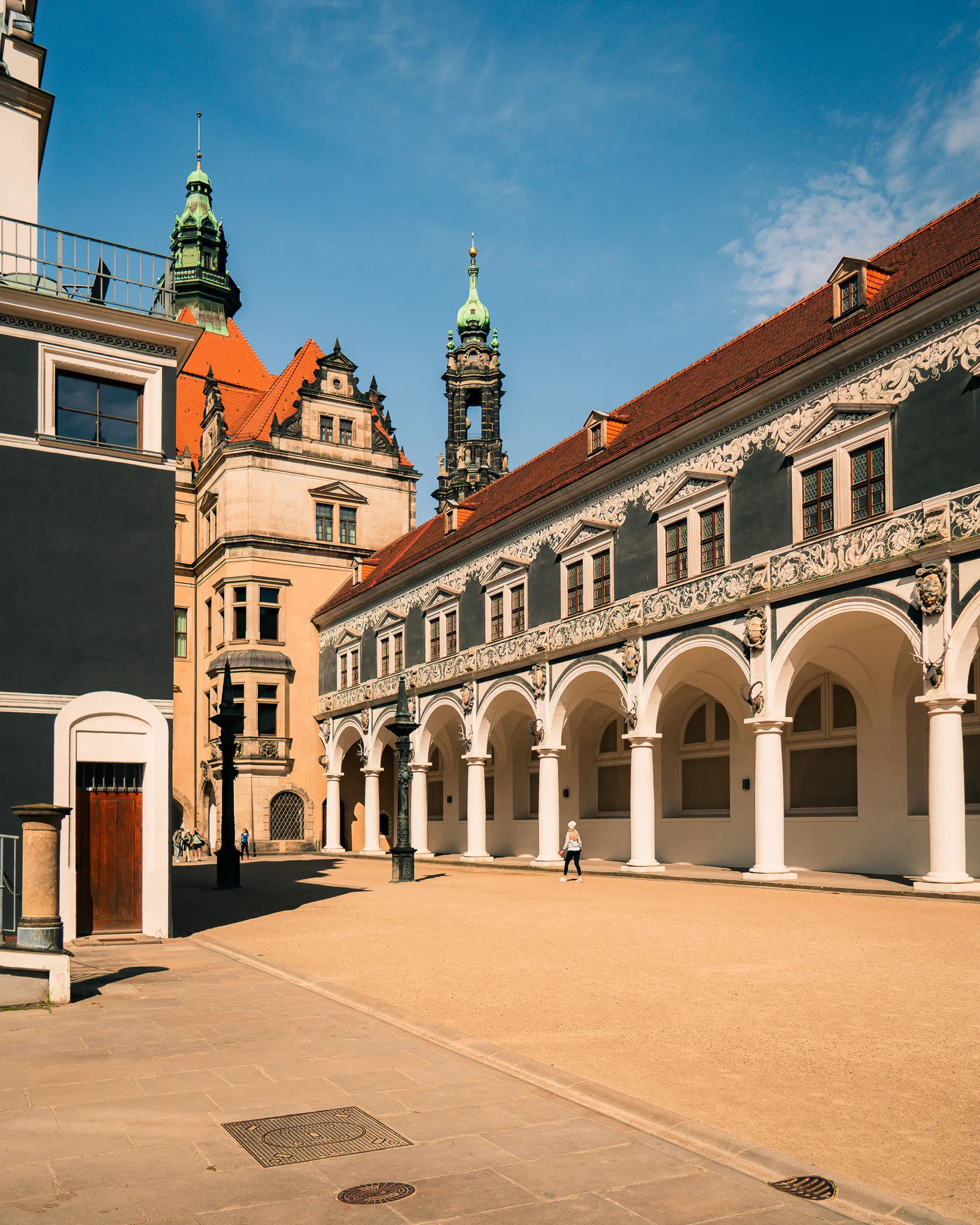 Spacerowicze w wąskiej uliczce przy Zamku Królewskim w Dreźnie – widok na bogato zdobioną fasadę z czerwonym dachem