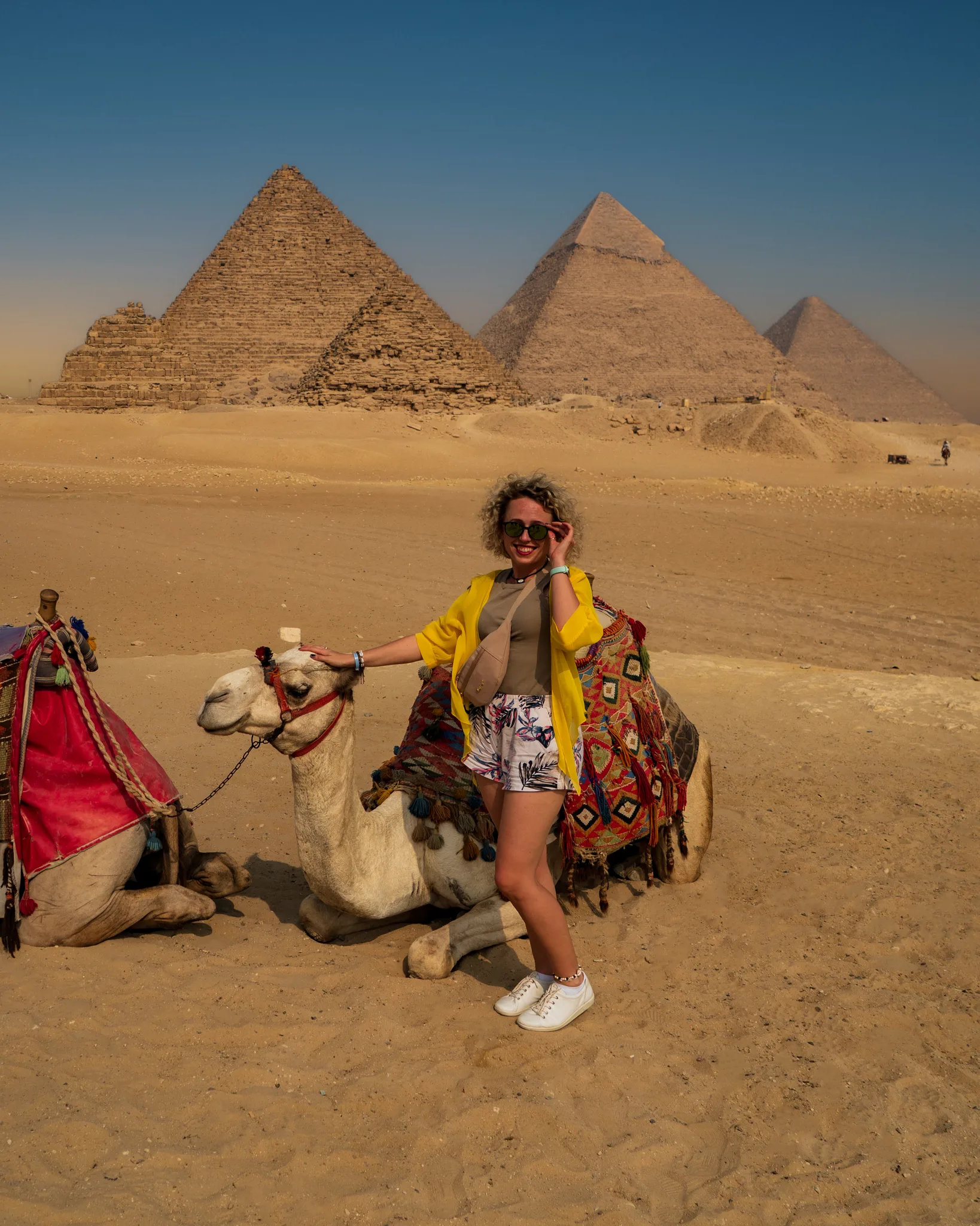 Turystka siedząca przy udekorowanym wielbłądzie na pustyni w Gizie; w tle widoczne piramidy Cheopsa, Chefrena i Menkaure.