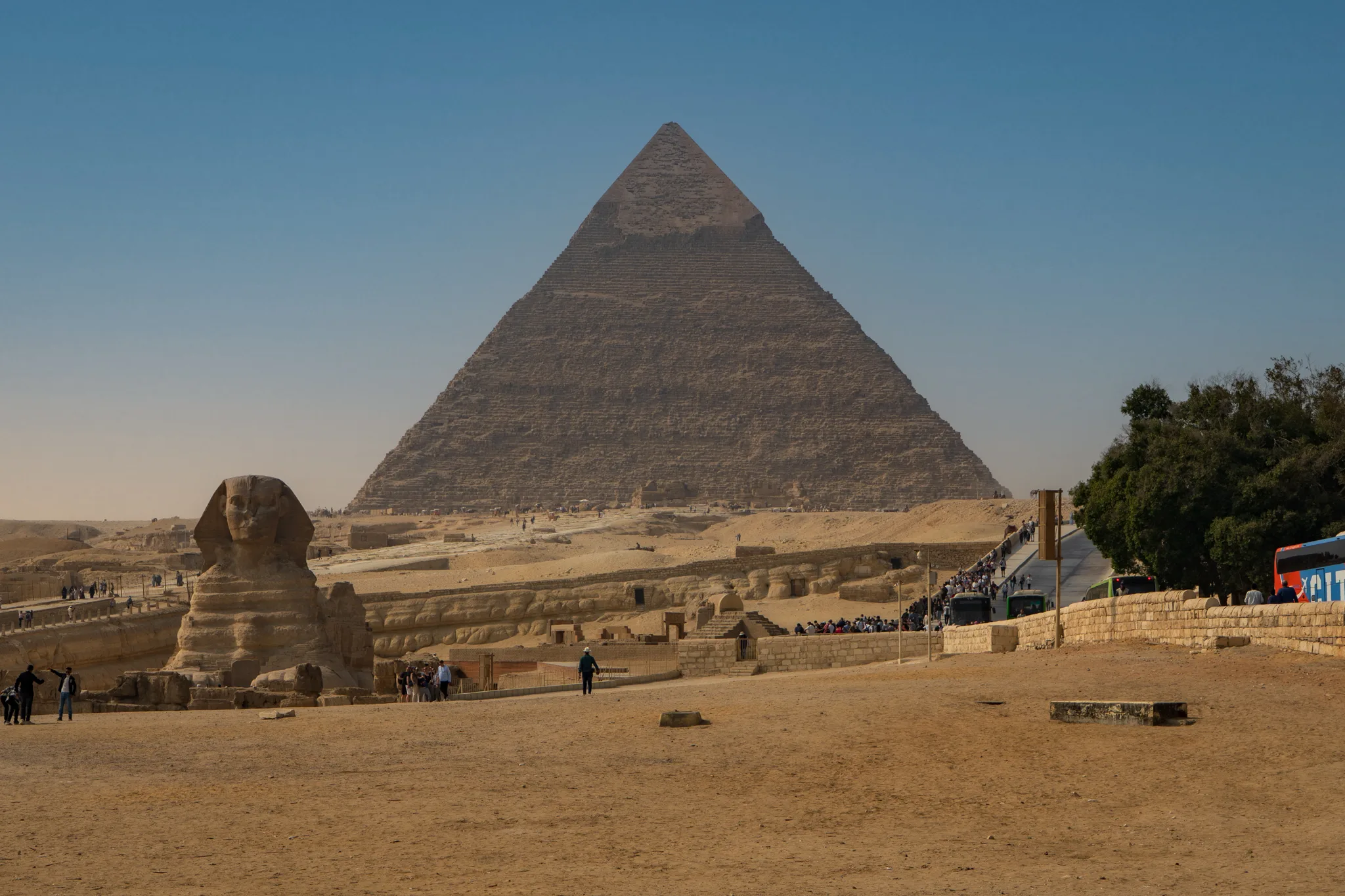 Panorama płaskowyżu Gizy z widokiem na Wielkiego Sfinksa i Piramidę Chefrena; na pierwszym planie pustynny teren i turyści.