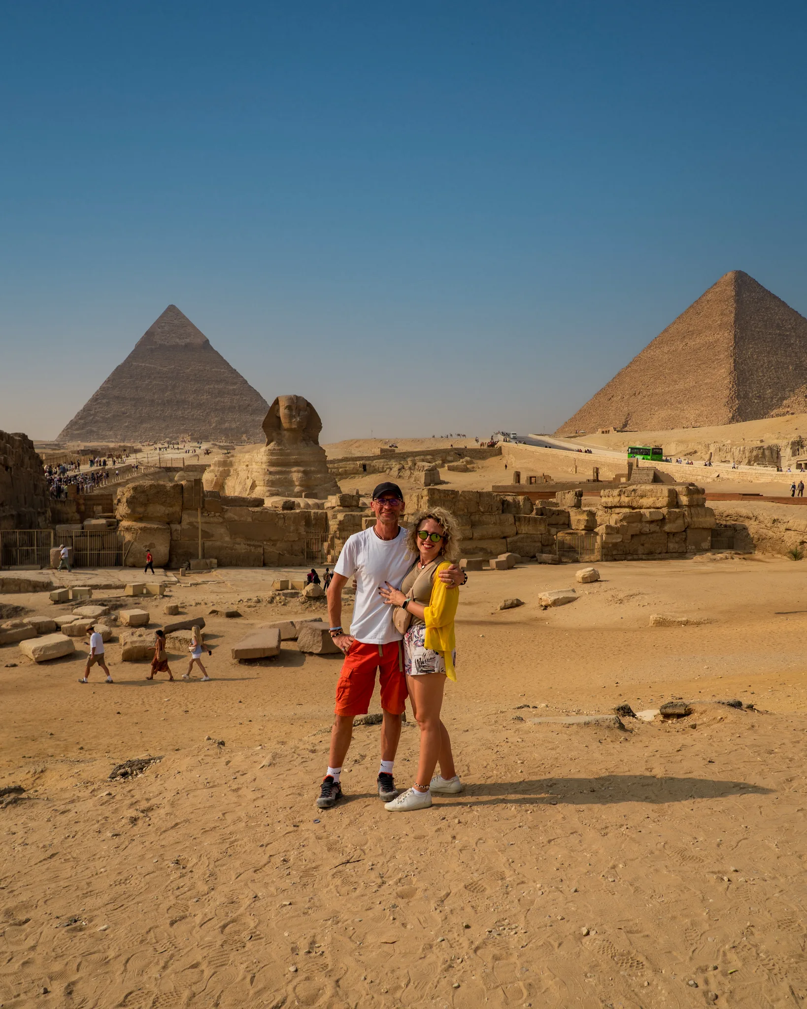 Panorama płaskowyżu Gizy z piramidami i Sfinksem; na pierwszym planie para turystów stojących na pustynnym terenie, z monumentalnymi budowlami w tle.