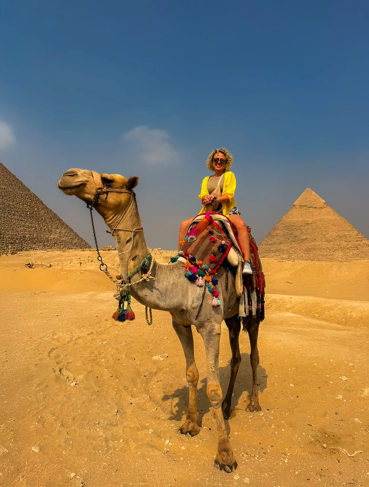Turystka siedząca na wielbłądzie na pustyni w Gizie; za nią dwie piramidy widoczne na horyzoncie.