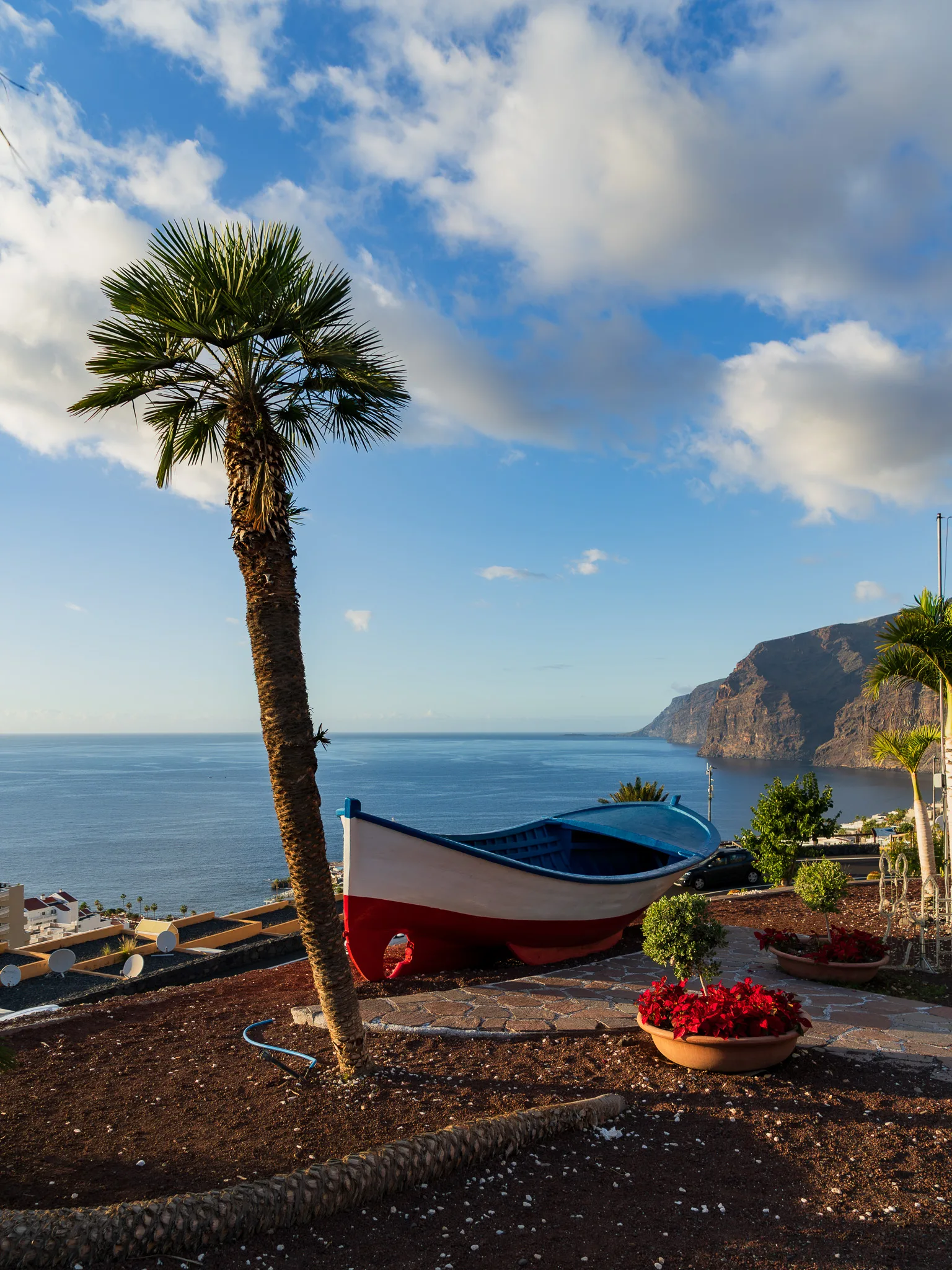 Łódź z widokiem na klify Los Gigantes – kolorowa, tradycyjna łódź ustawiona na punkcie widokowym nad oceanem, z palmą na pierwszym planie i monumentalnymi klifami opadającymi do Atlantyku w tle, w ciepłym świetle popołudnia.