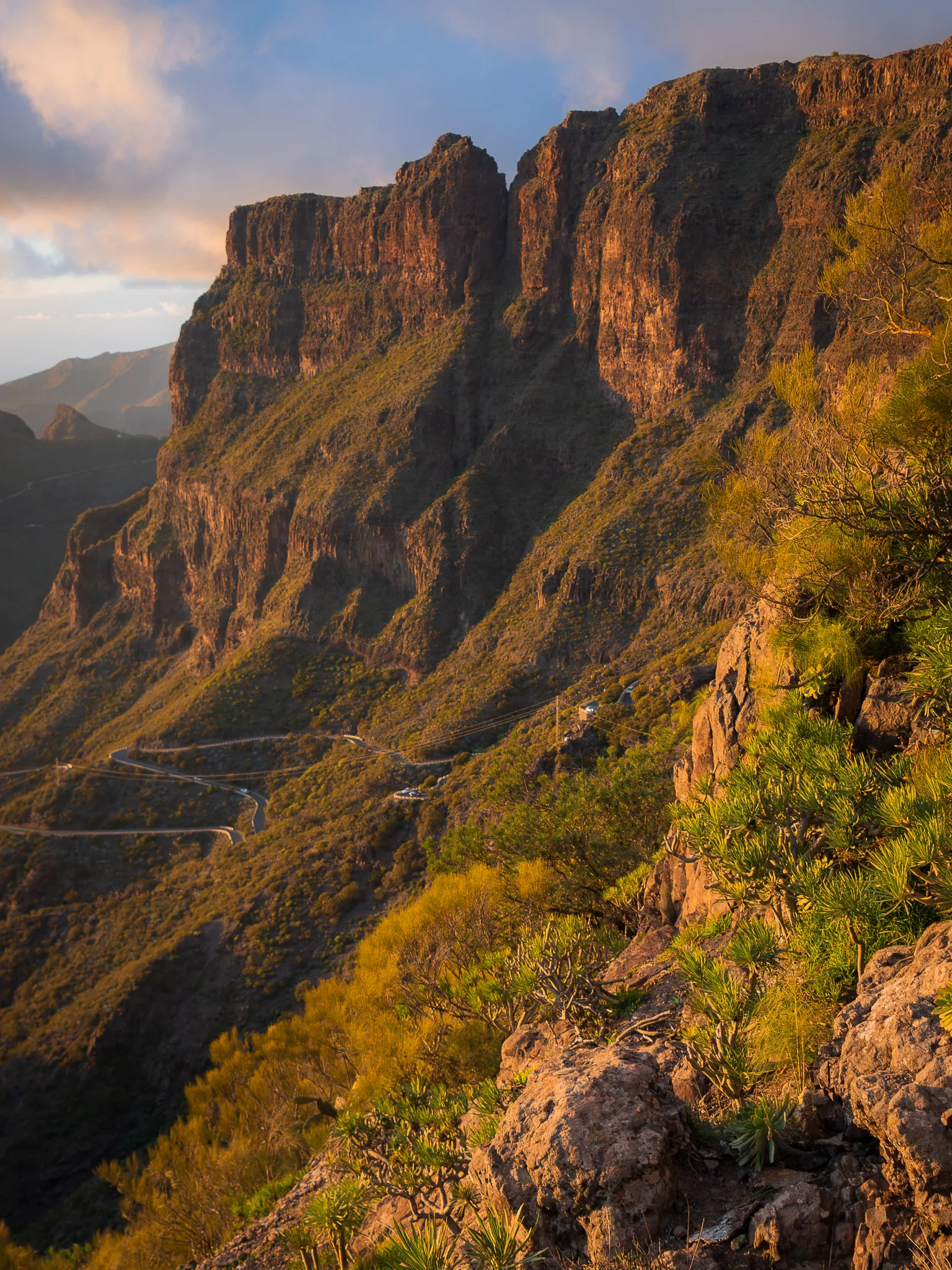 Klify wąwozu Masca o zachodzie słońca – monumentalne ściany skalne i serpentyny drogi wijącej się w dole, podkreślone złotym światłem i dramatycznym krajobrazem gór TENERYFA