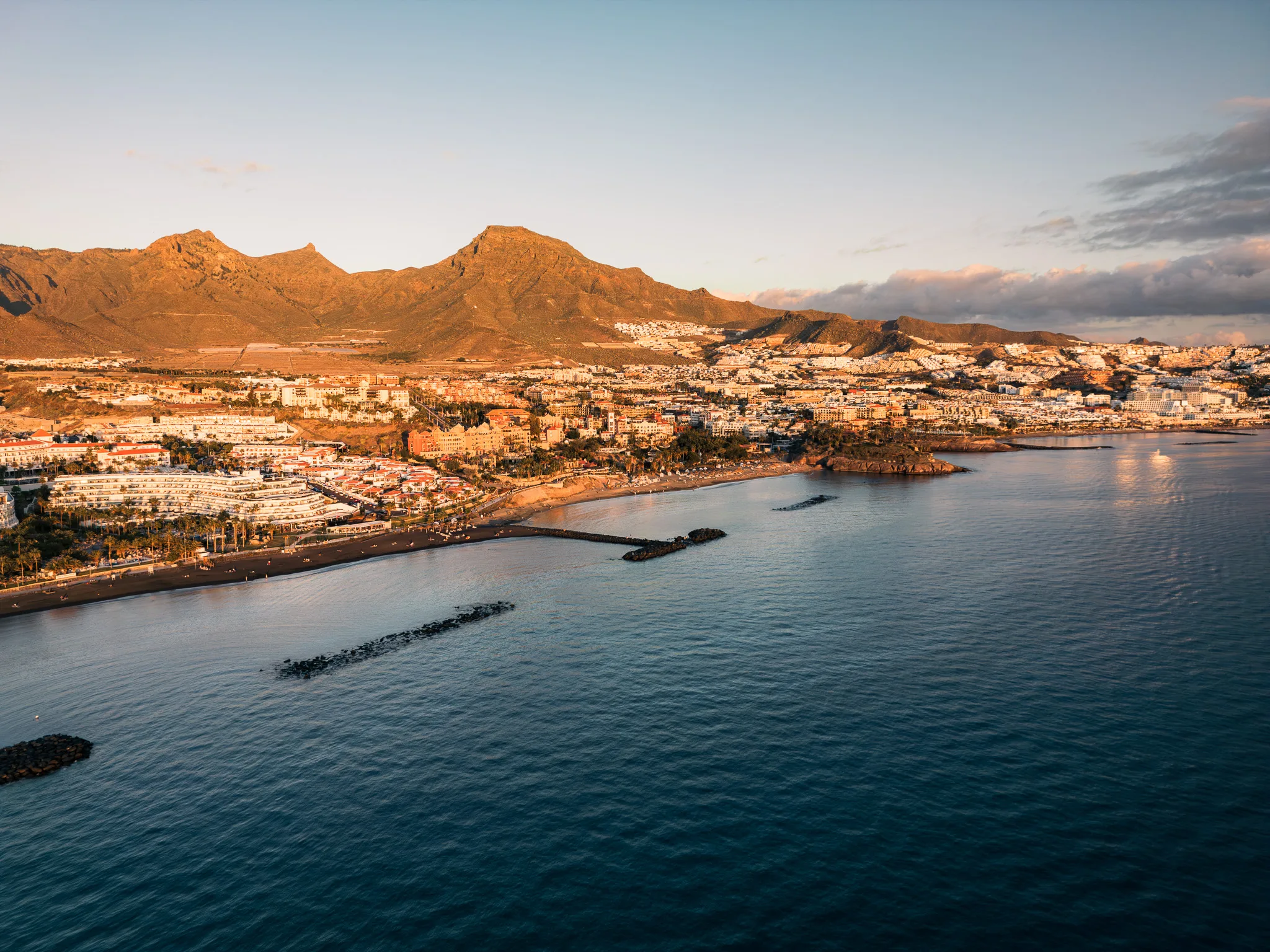 Widok z drona na skalisty klif i nadmorskie miasteczko o zachodzie słońca, z jasną zabudową, oceanem i górami w tle.