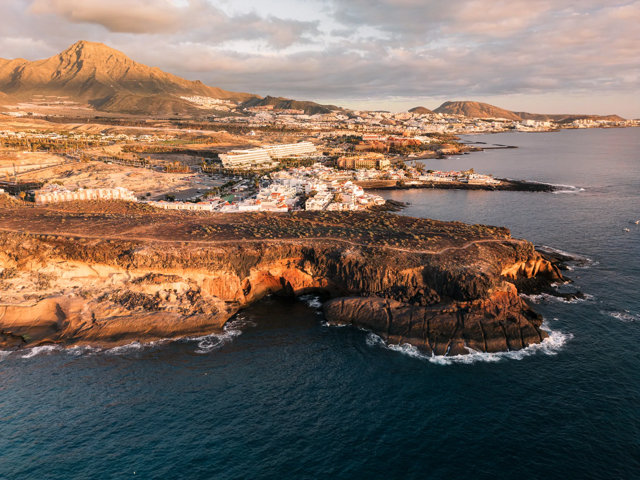 Widok z drona na skalisty klif i nadmorskie miasteczko o zachodzie słońca, z jasną zabudową, oceanem i górami w tle.