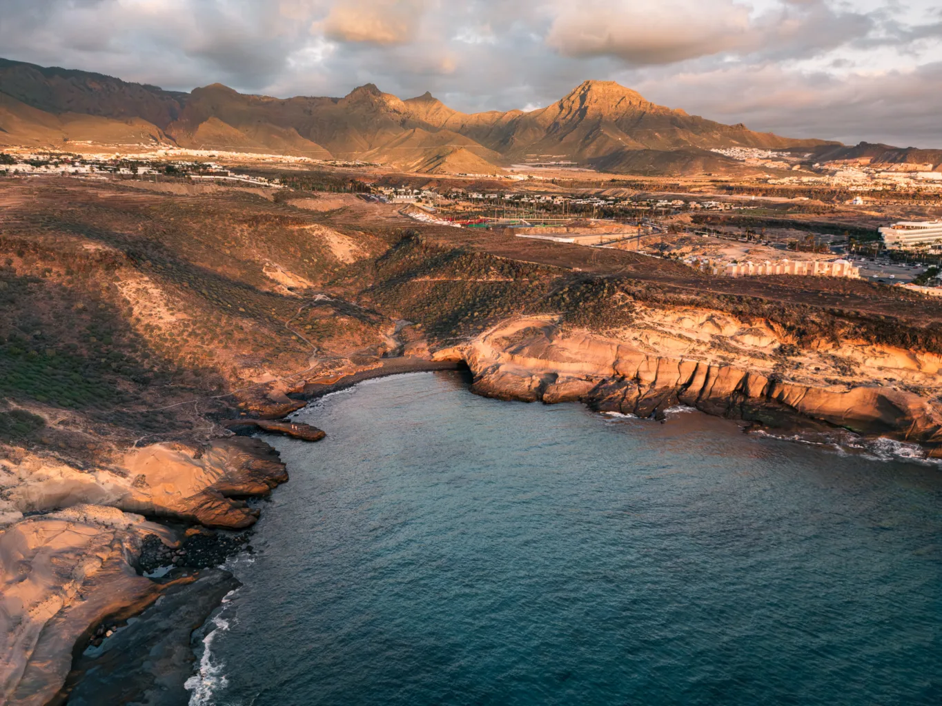 Widok z drona na skalisty klif i nadmorskie miasteczko o zachodzie słońca, z jasną zabudową, oceanem i górami w tle.