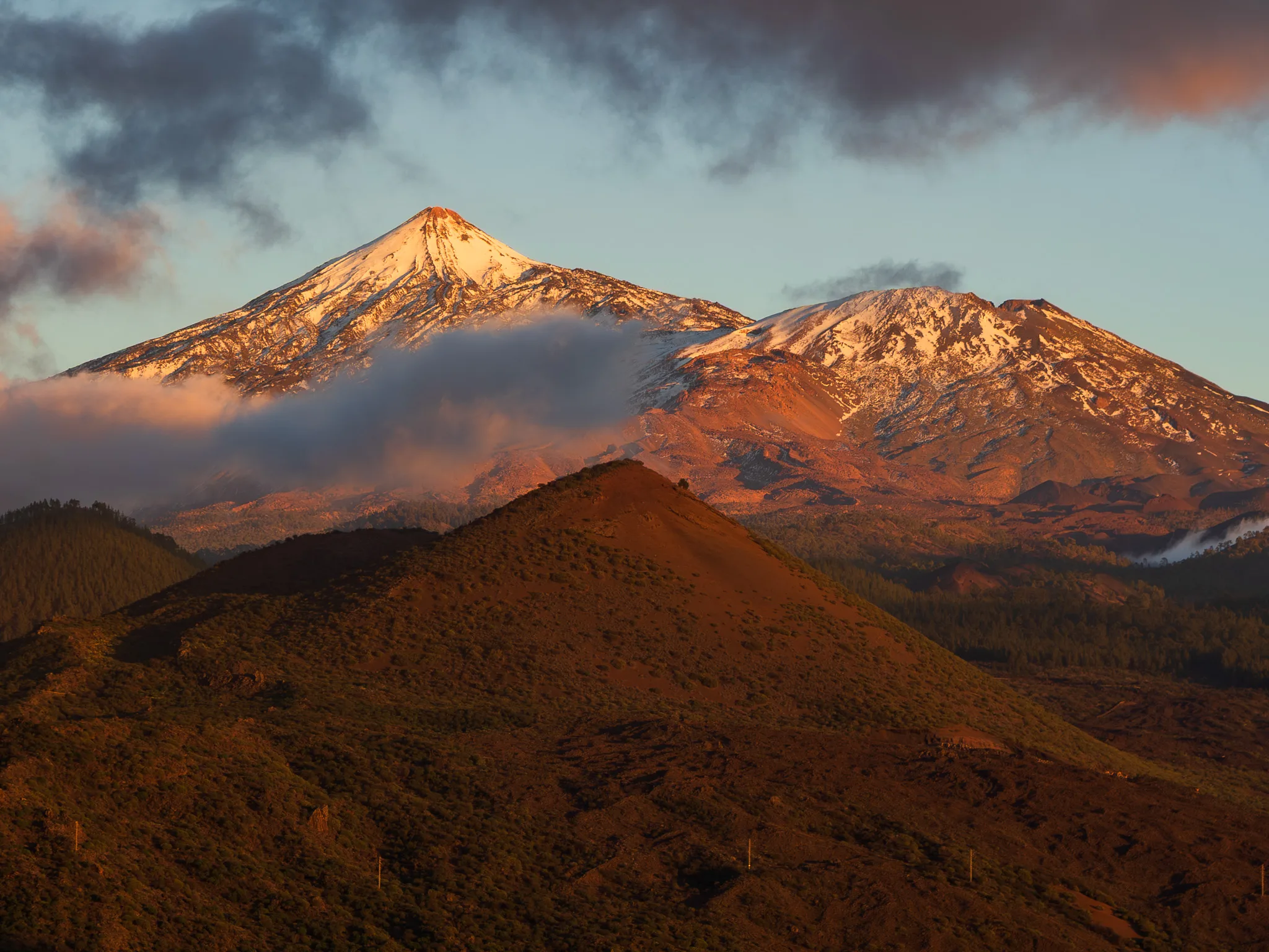 Wulkan Teide o zachodzie słońca – ośnieżony szczyt Teide skąpany w ciepłym, złotym świetle, z niskimi chmurami sunącymi po zboczach i kontrastem między jasnym śniegiem a ciemnymi, wulkanicznymi stokami Teneryfy.