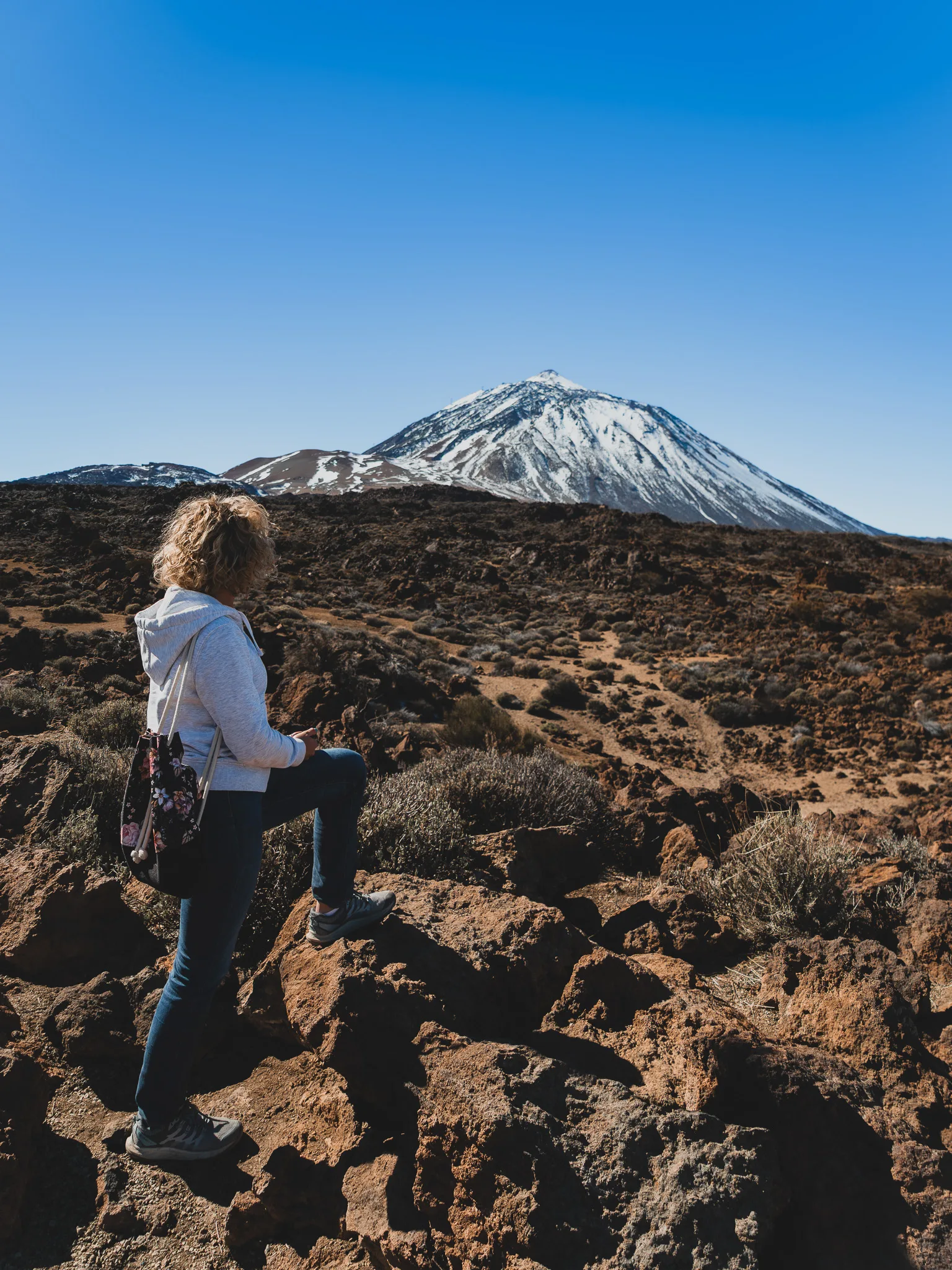 Widok na wulkan Teide z perspektywy szlaku – osoba stojąca na lawowych skałach, przed nią surowy, wulkaniczny krajobraz i ośnieżony szczyt Teide na tle intensywnie niebieskiego nieba.