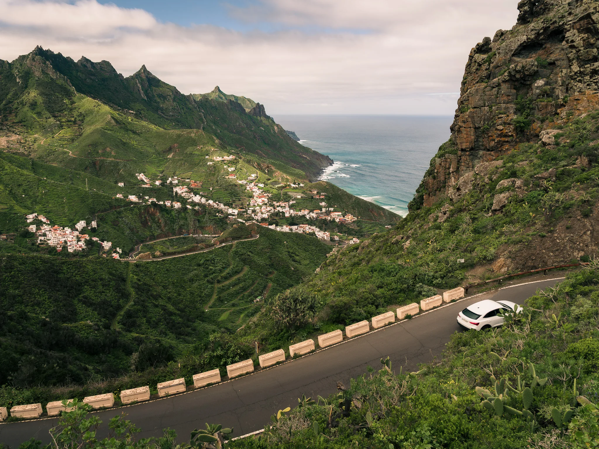 Kręta droga w górach Anaga na Teneryfie, białe auto na serpentynie nad zieloną doliną i oceanem w tle.