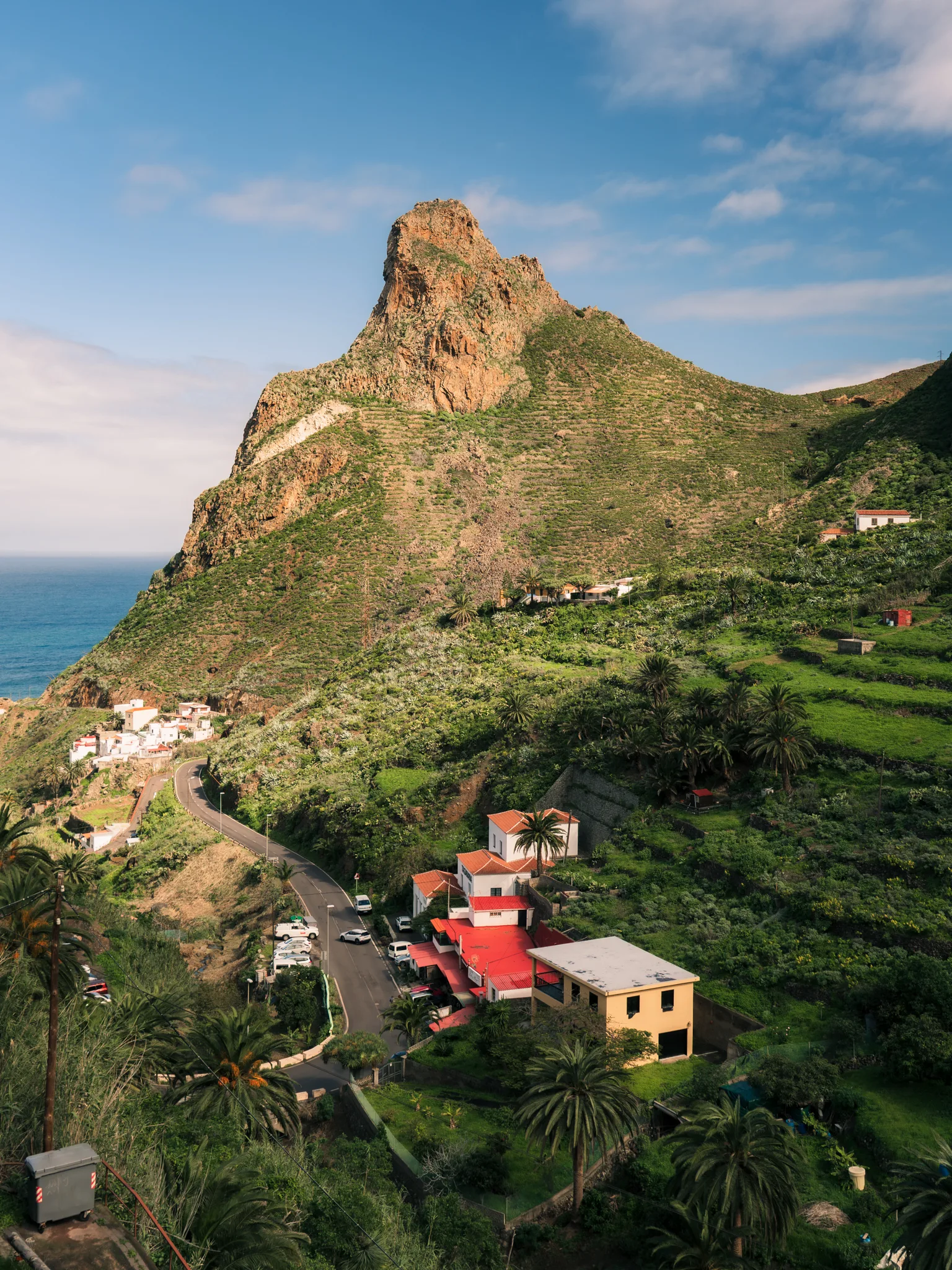 Zielona dolina północnej Teneryfy z charakterystyczną skalną formacją, krętą drogą i zabudową wśród palm oraz tarasów rolniczych.