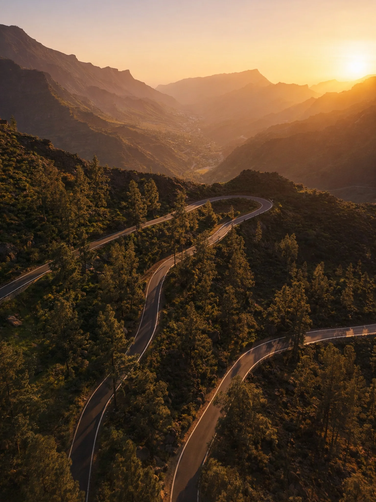 Kręta górska droga z serpentynami na Gran Canarii podczas delikatnego zachodu słońca, miękkie cienie, złota godzina z prawej strony, zielone zbocza i warstwowe pasma gór w tle.
