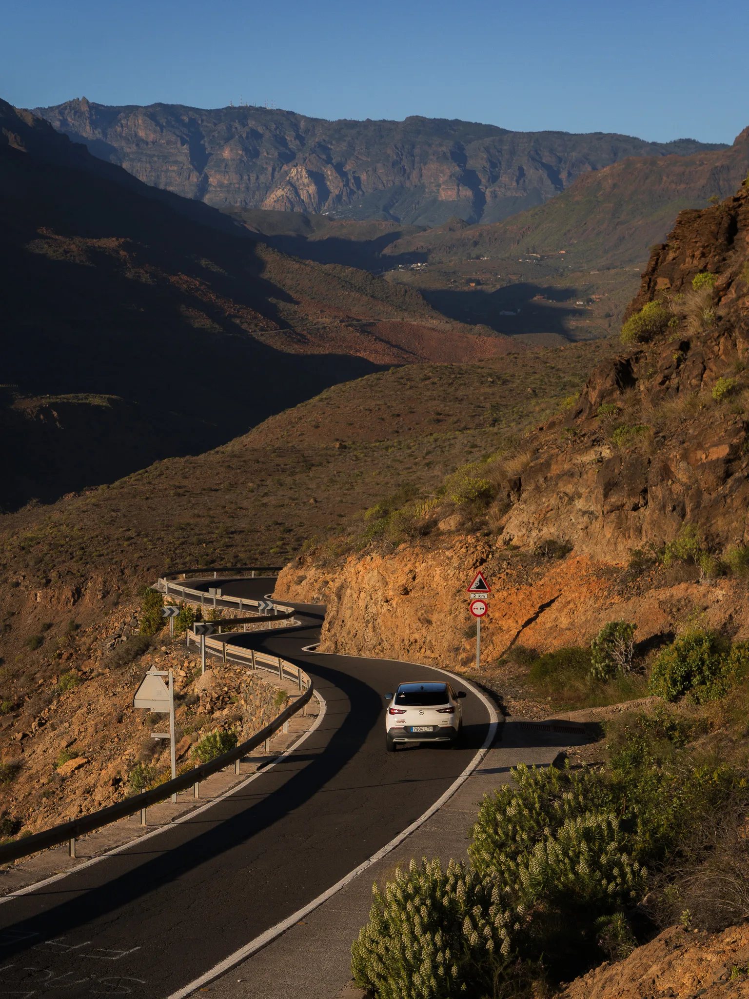 Kręta górska droga z białym autem na serpentynach, skaliste zbocza i rozległa dolina w tle - Gran Canaria