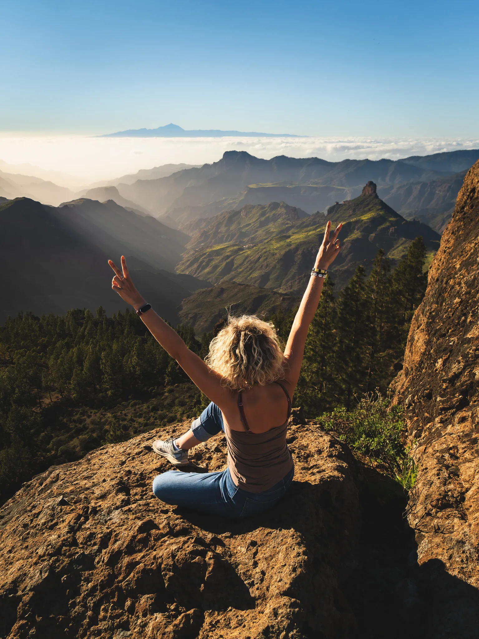 Gran Canaria: kobieta siedzi na skale i pokazuje znak zwycięstwa, panorama gór i morze chmur o złotej godzinie