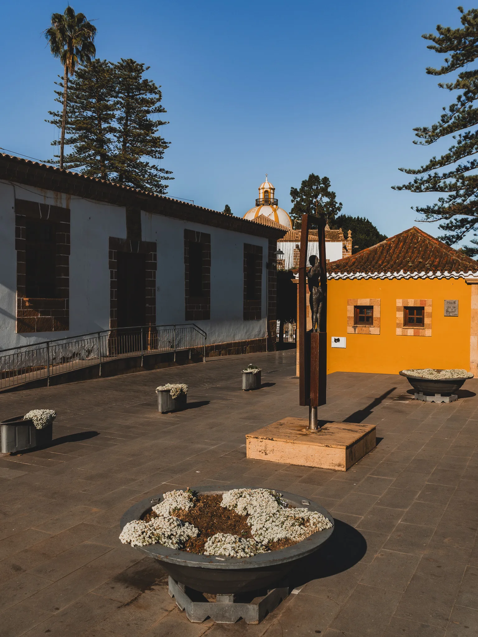 Gran Canaria: Teror – spokojny plac z żółtym budynkiem i rzeźbą, w tle kopuła bazyliki