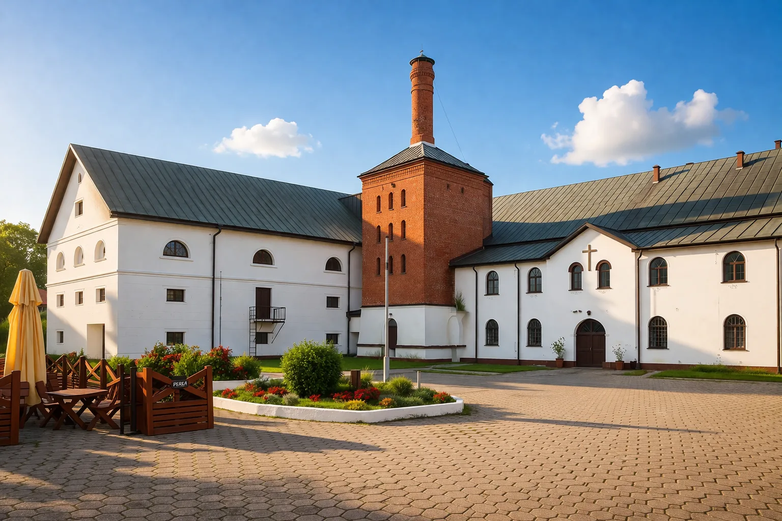 Zabytkowy browar w Zwierzyńcu z charakterystyczną ceglaną wieżą w słoneczny poranek, na tle czystego nieba z delikatnymi chmurami, otoczony zadbaną zielenią i brukowanym dziedzińcem